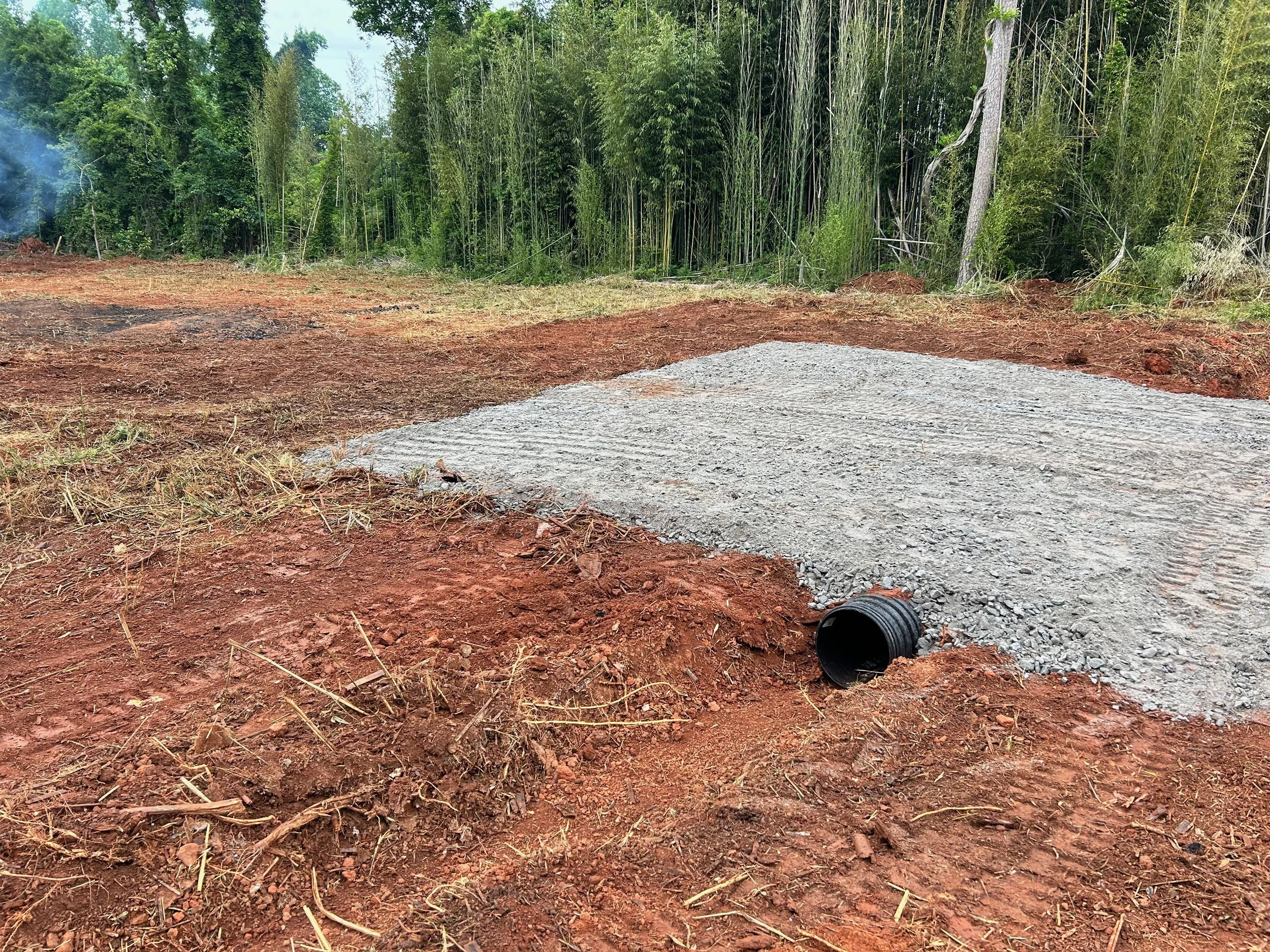 Excavated ground with a gravel pad, black drainage pipe, nearby forest with tall trees and bamboo, and some smoke in the distance.