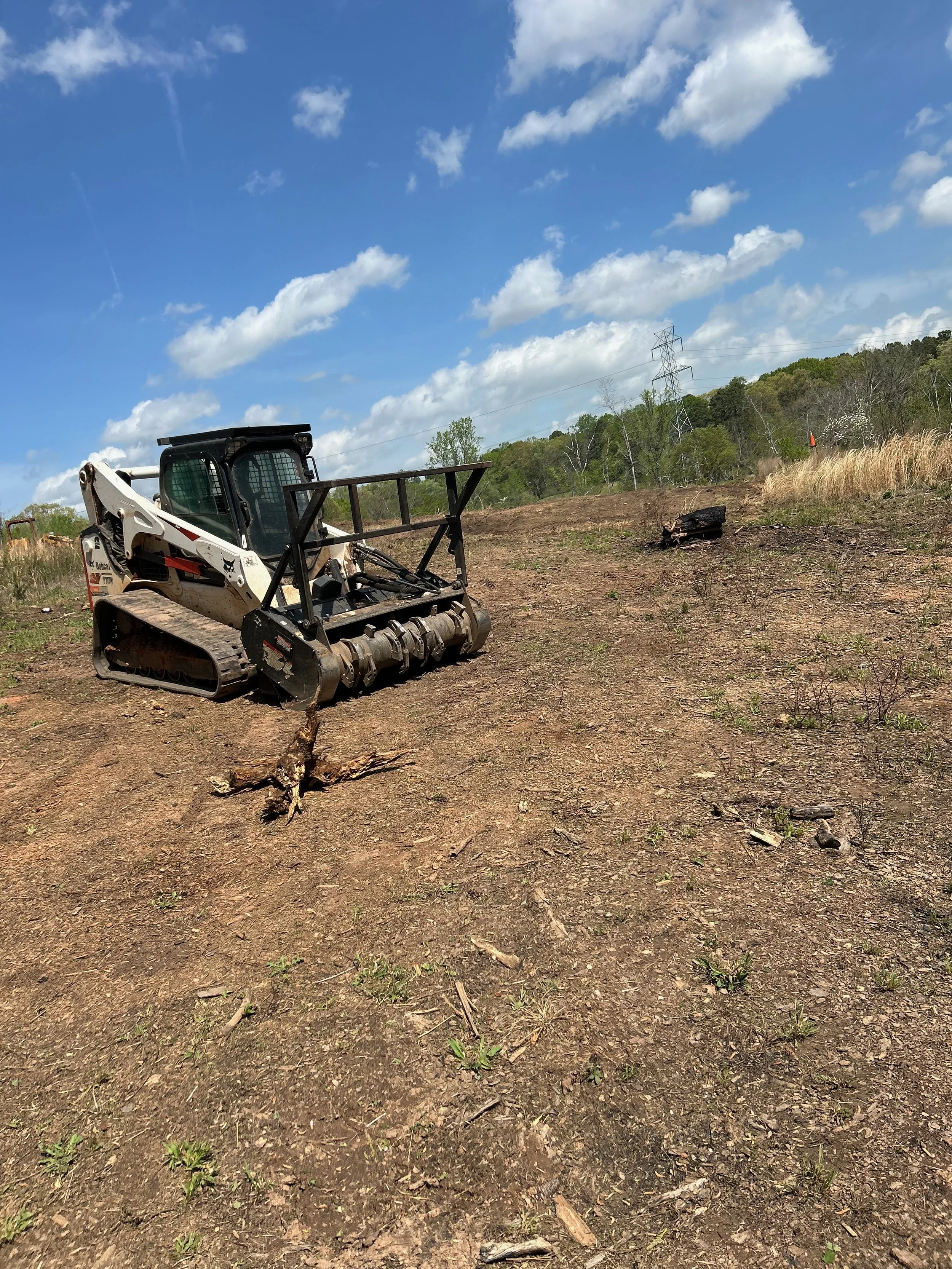 A skid-steer loader on a cleared dirt field with some small plants, grass, and a tree stump, under a partly cloudy sky.