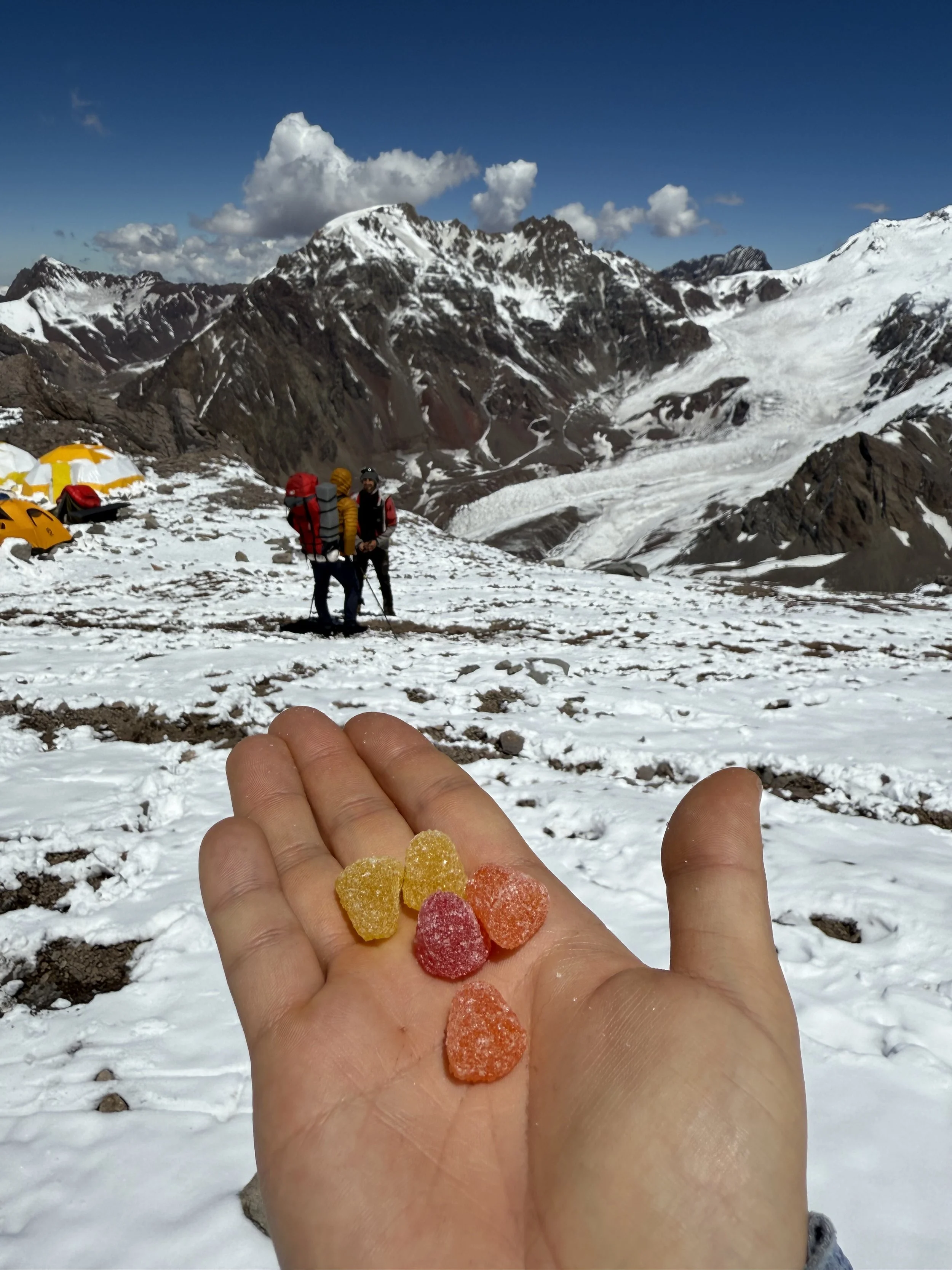 A hand holding colorful gummy candies with a snow-covered mountain landscape and two climbers in the background.