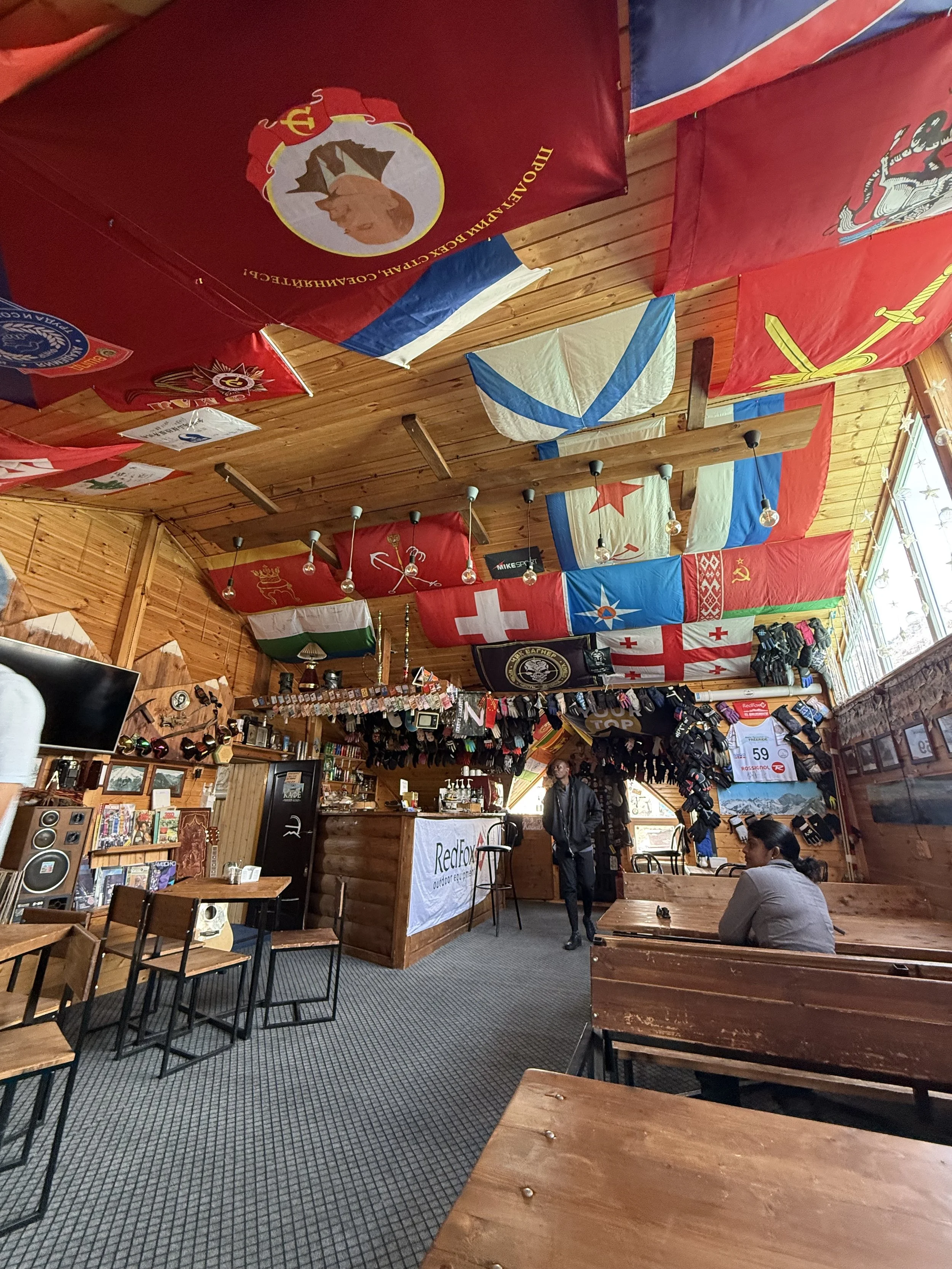 Interior of a cozy wooden cafe decorated with various flags hanging from the ceiling, including military, national, and historical flags, with a counter, tables, chairs, and a few customers.