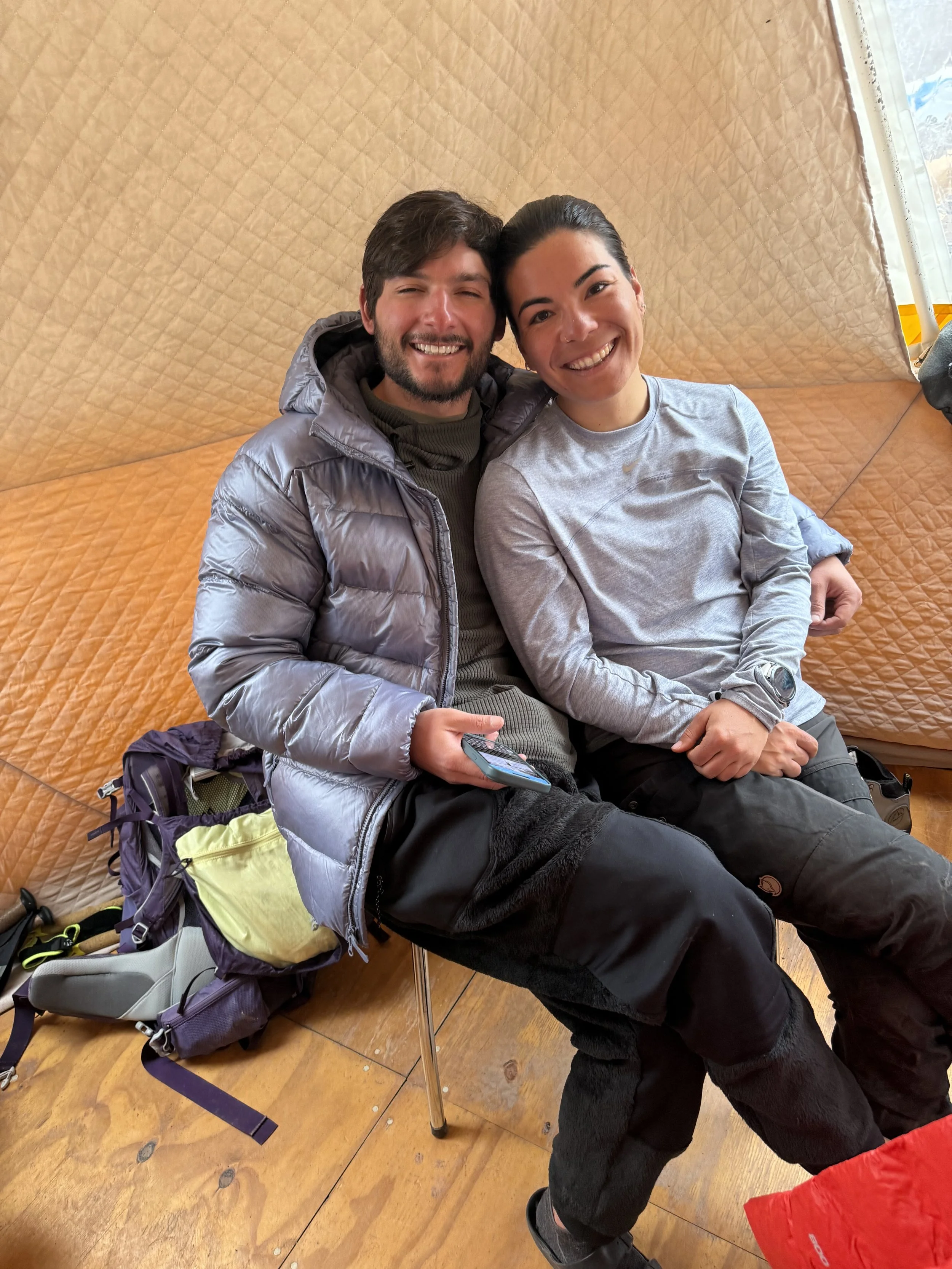 Two people sitting close together inside a padded tent, smiling at the camera. One is wearing a puffy gray jacket and the other a light gray long-sleeve shirt. There is a backpack and gear on the floor beside them.