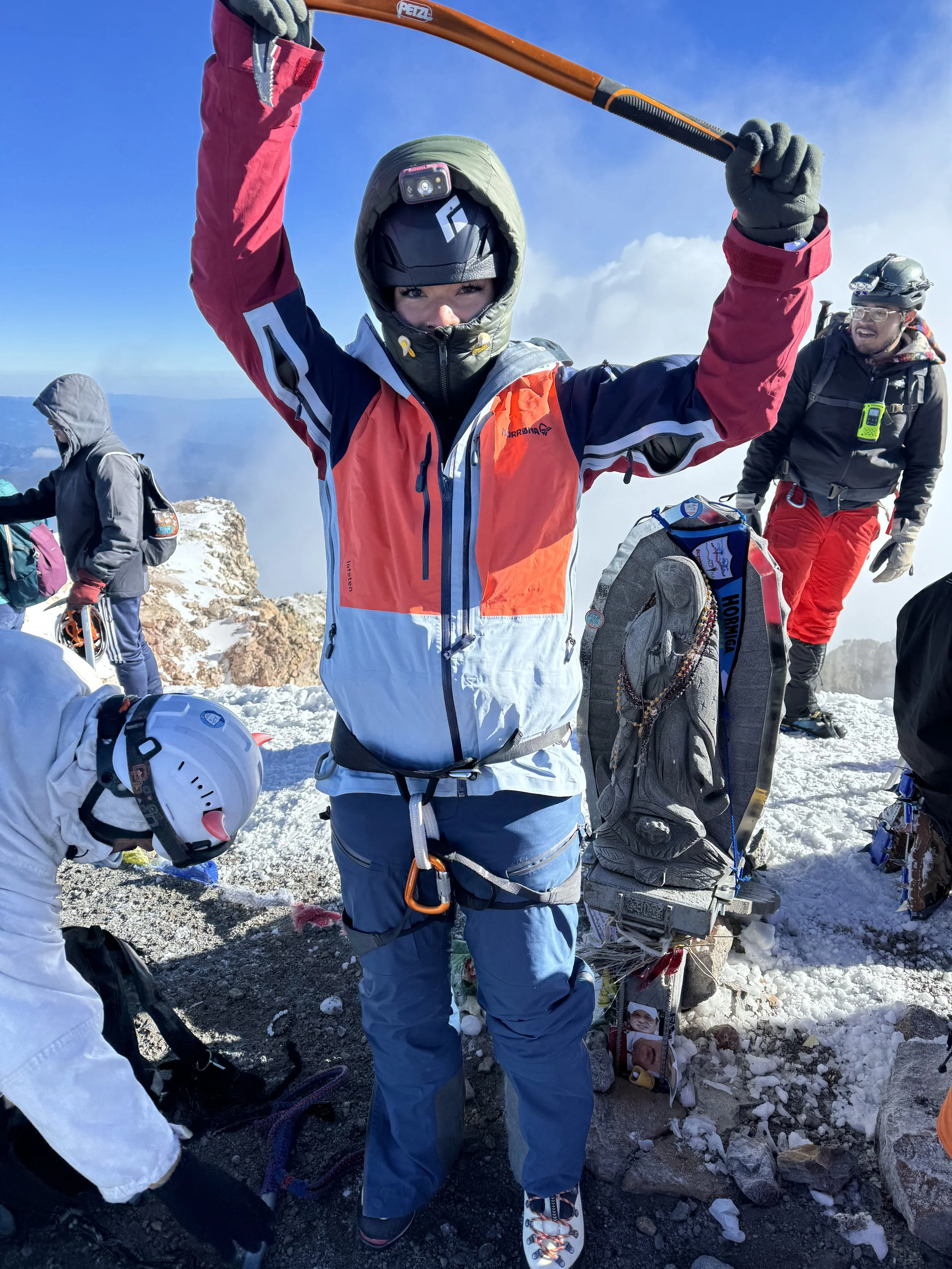 A mountain climber at the summit, holding an ice axe above their head, surrounded by snow and ice, with other climbers in the background.