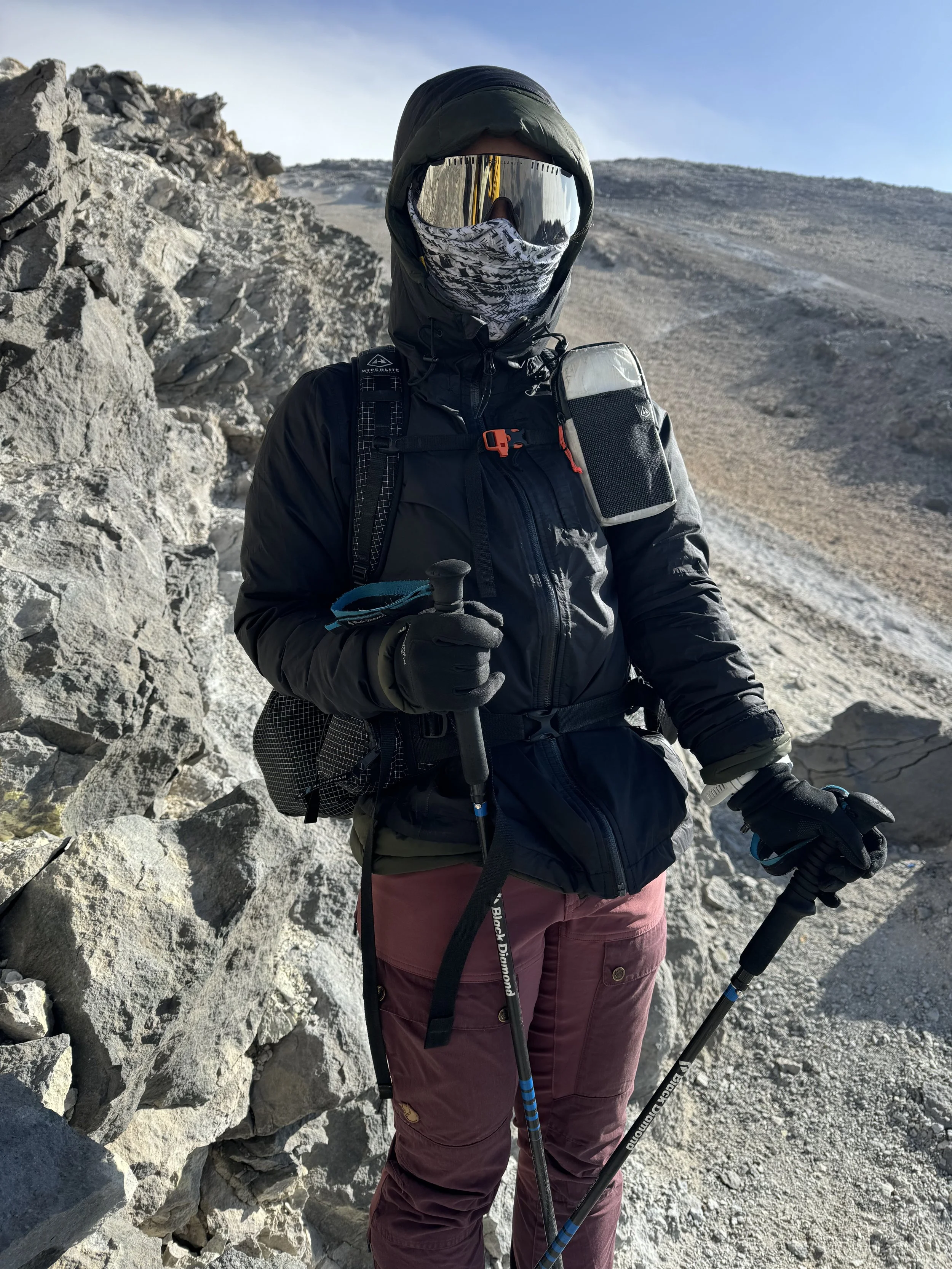 A person dressed in outdoor hiking gear, including a jacket, cargo pants, and gloves, stands with hiking poles in a rocky mountain landscape with a clear sky.