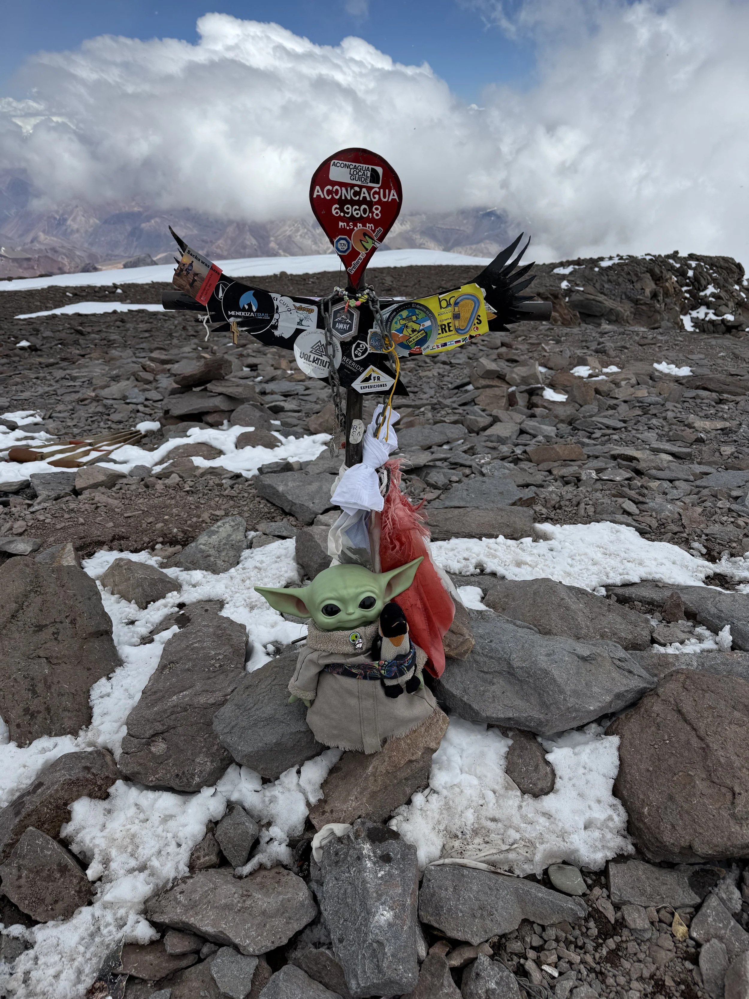 A summit marker at Aconcagua, indicating an elevation of 6,960.8 meters, surrounded by rocks and snow on a mountain landscape with clouds and distant mountain peaks.