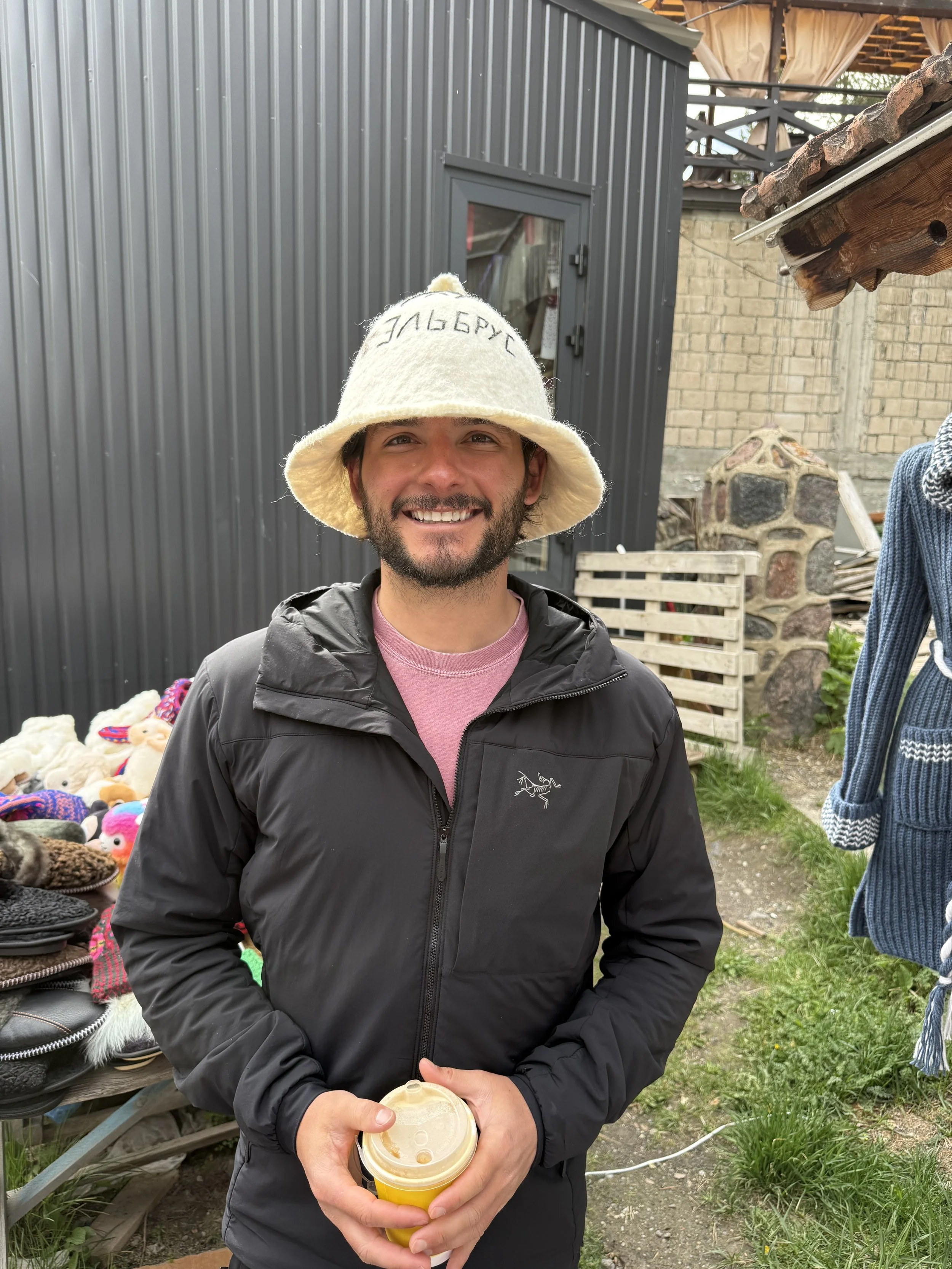 A smiling man holding a coffee cup outdoors at a market or craft fair, wearing a black jacket and a cream-colored felt hat with embroidery, with various items displayed on tables in the background.