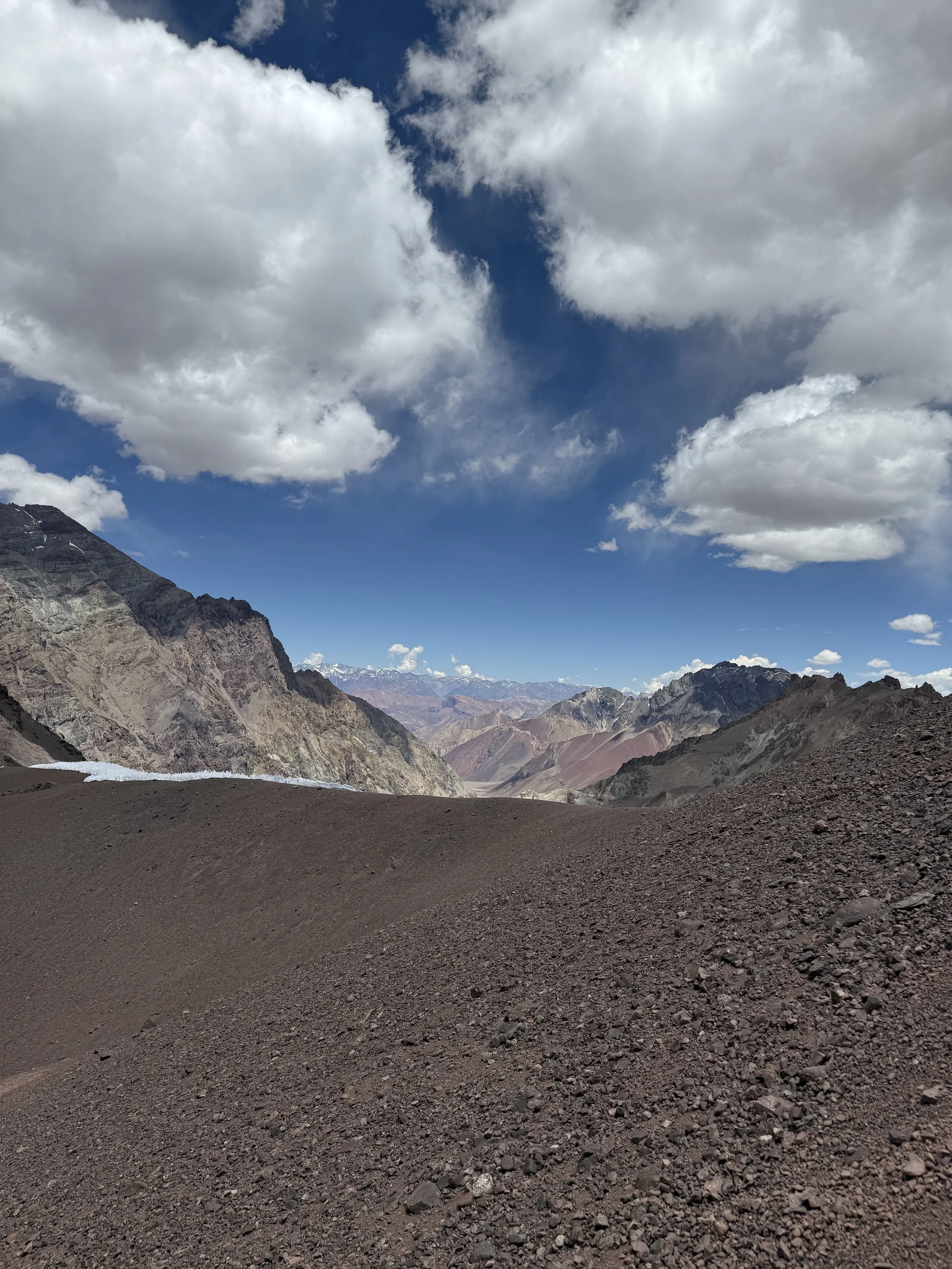 View of a rugged mountain landscape with rocky terrain in the foreground, colorful mountains in the distance, and a partly cloudy sky overhead.
