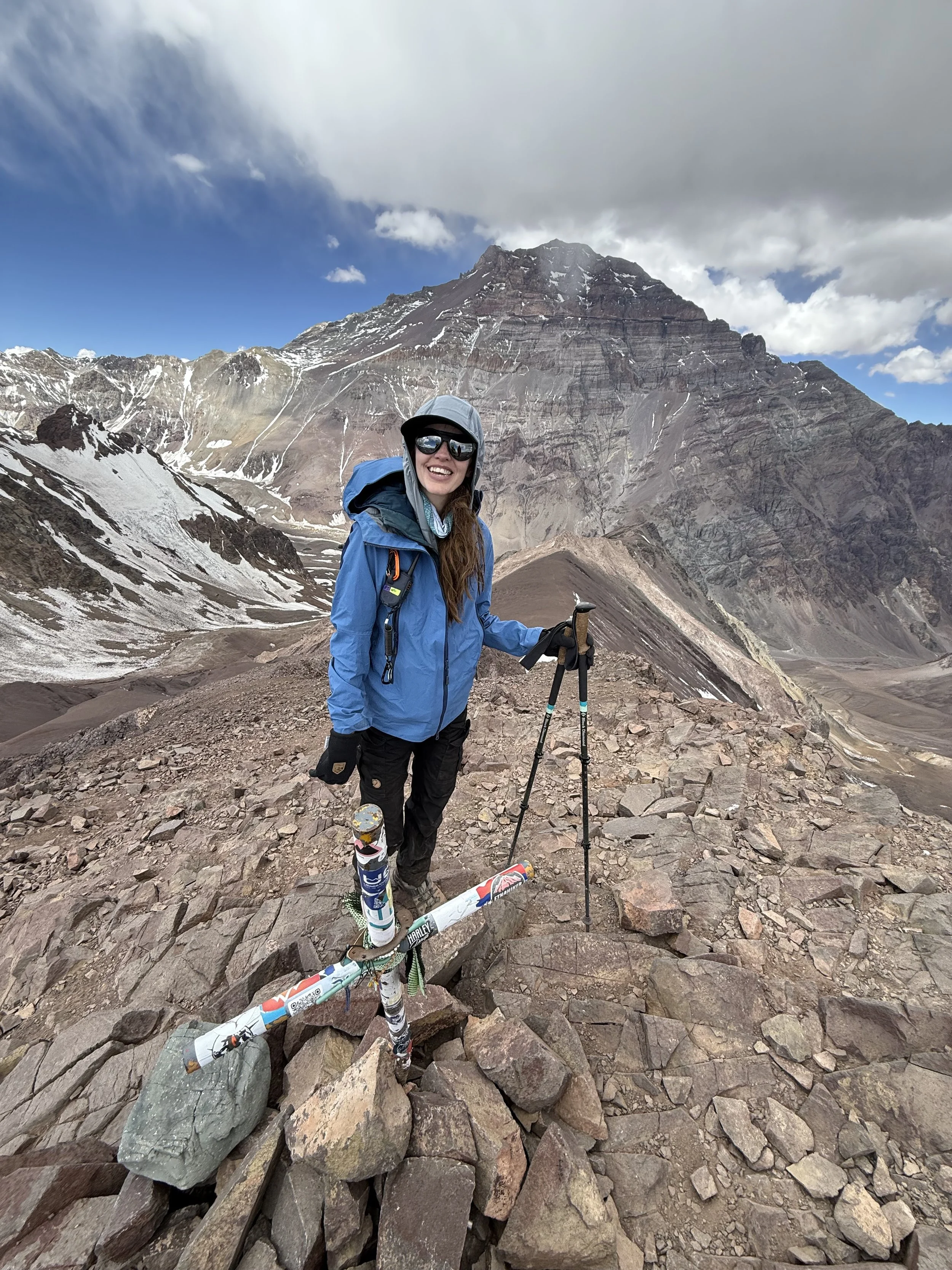 A woman standing on rocky terrain in a mountainous landscape, dressed in hiking gear with a blue jacket, black pants, helmet, and sunglasses, holding hiking poles, with snow-capped mountains and a cloudy sky in the background.