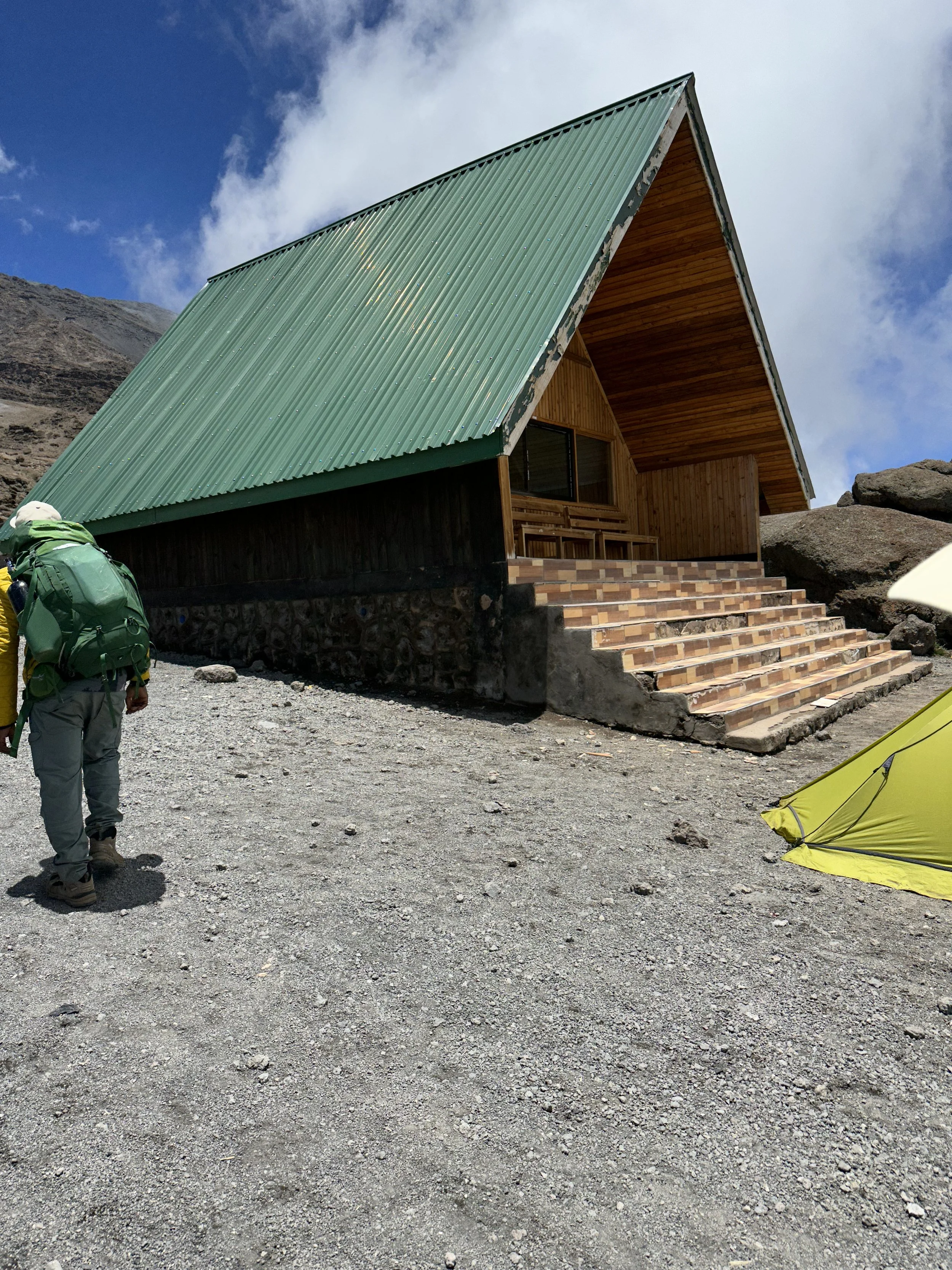 A small mountain cabin with a green metal roof and wooden walls, set against a cloudy sky. A person with a green backpack is walking toward the cabin, and a yellow tent is on the ground nearby.