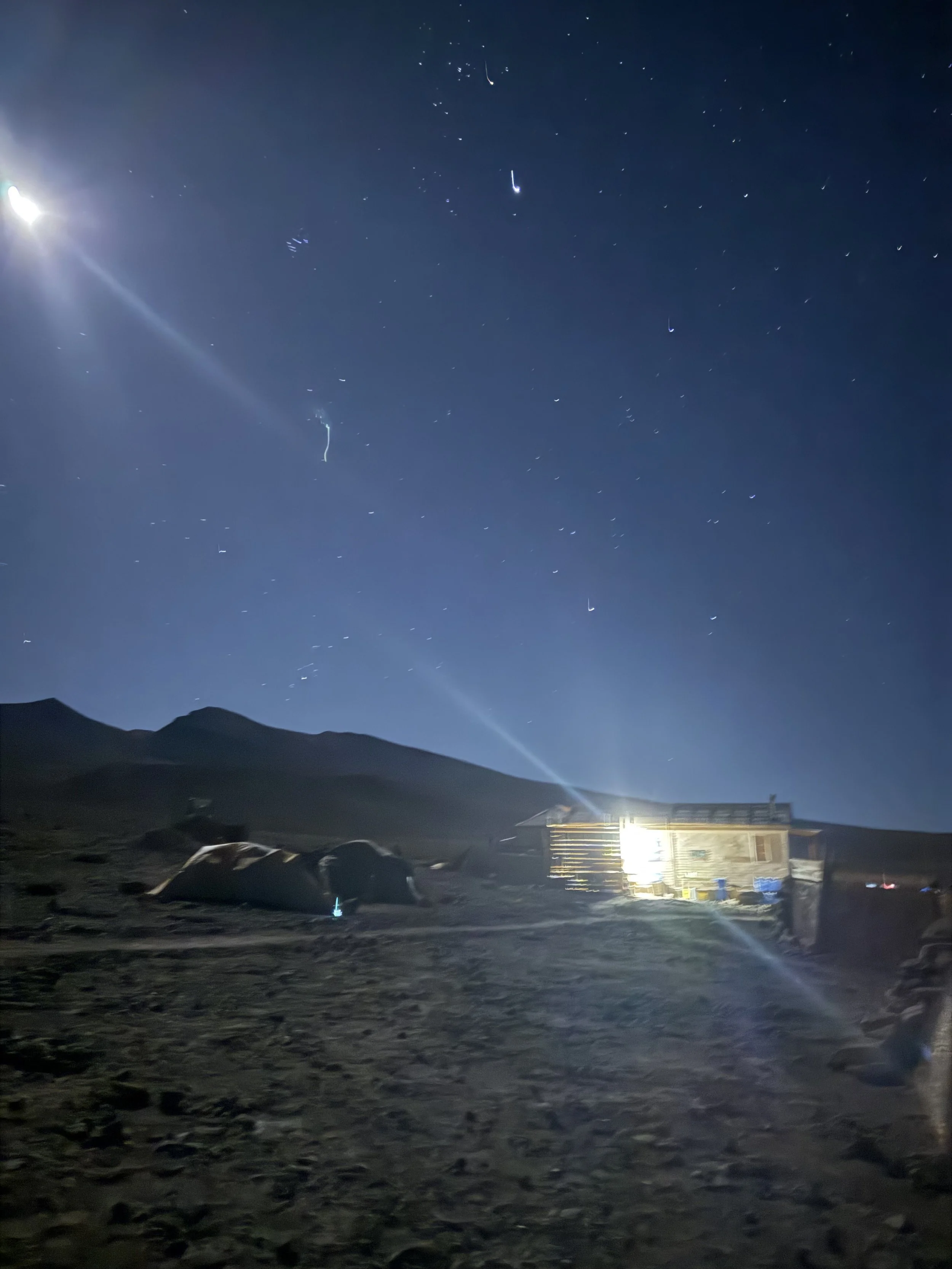 Nighttime scene of a desert landscape with a small structure emitting bright light, tents on the ground, distant mountains, and a starry sky.