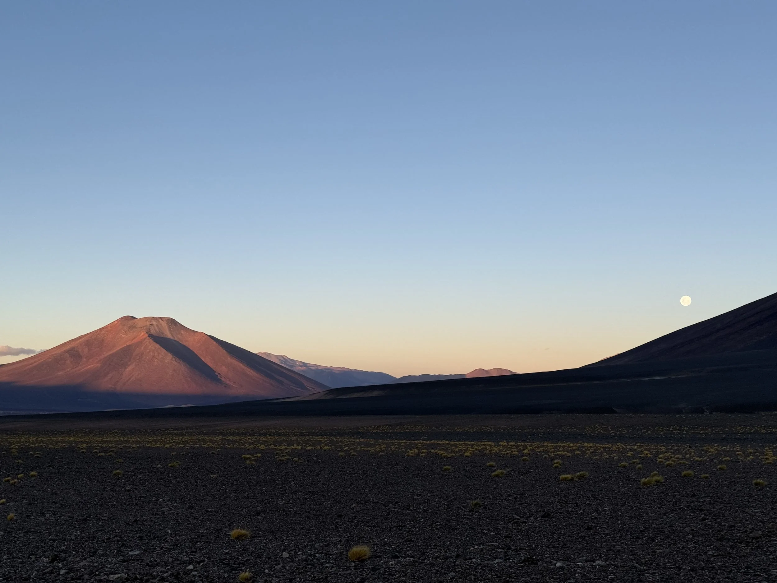 A desert landscape with mountains in the background under a clear sky, with a visible moon on the right side.
