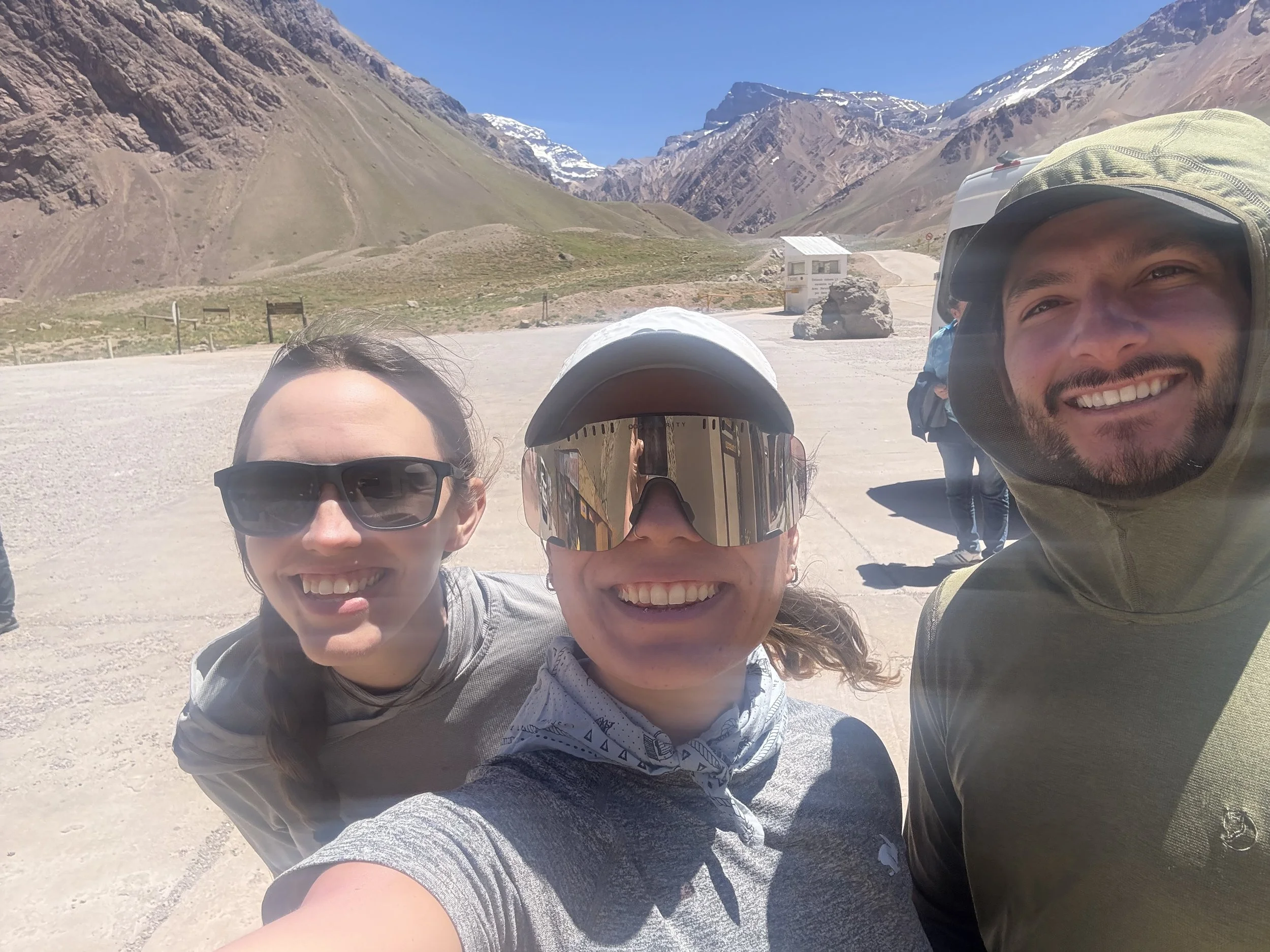 Three smiling friends taking a selfie outdoors in a mountainous area with a clear sky, snow-capped peaks, and a small white building in the background.