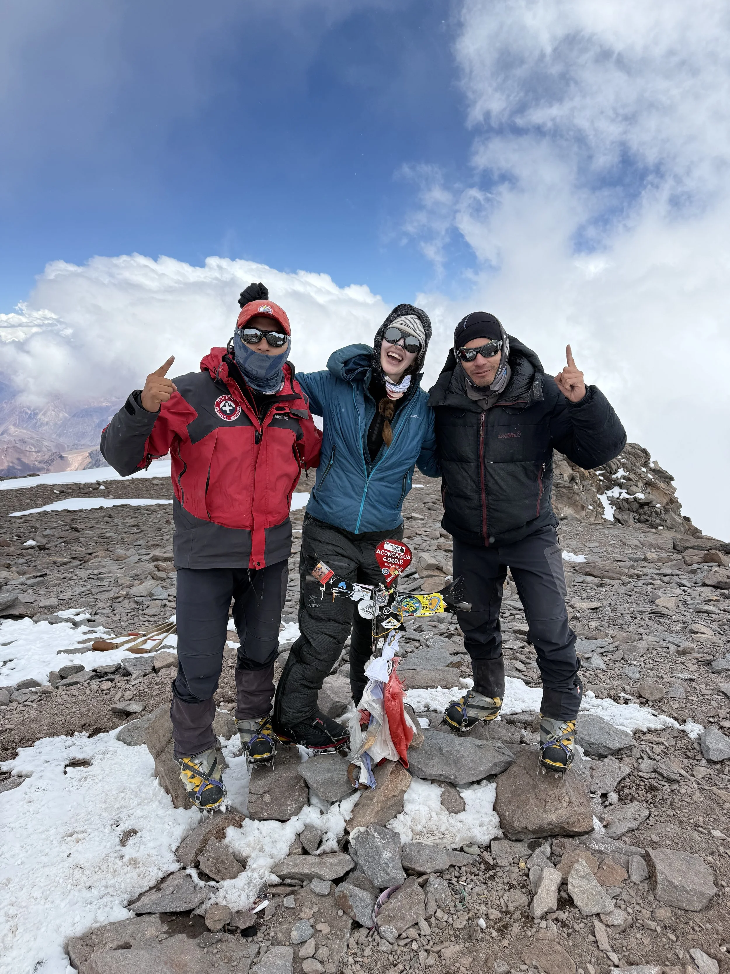 Three climbers standing on rocky mountaintop with snow and clouds, celebrating near a memorial with plaques and flags.