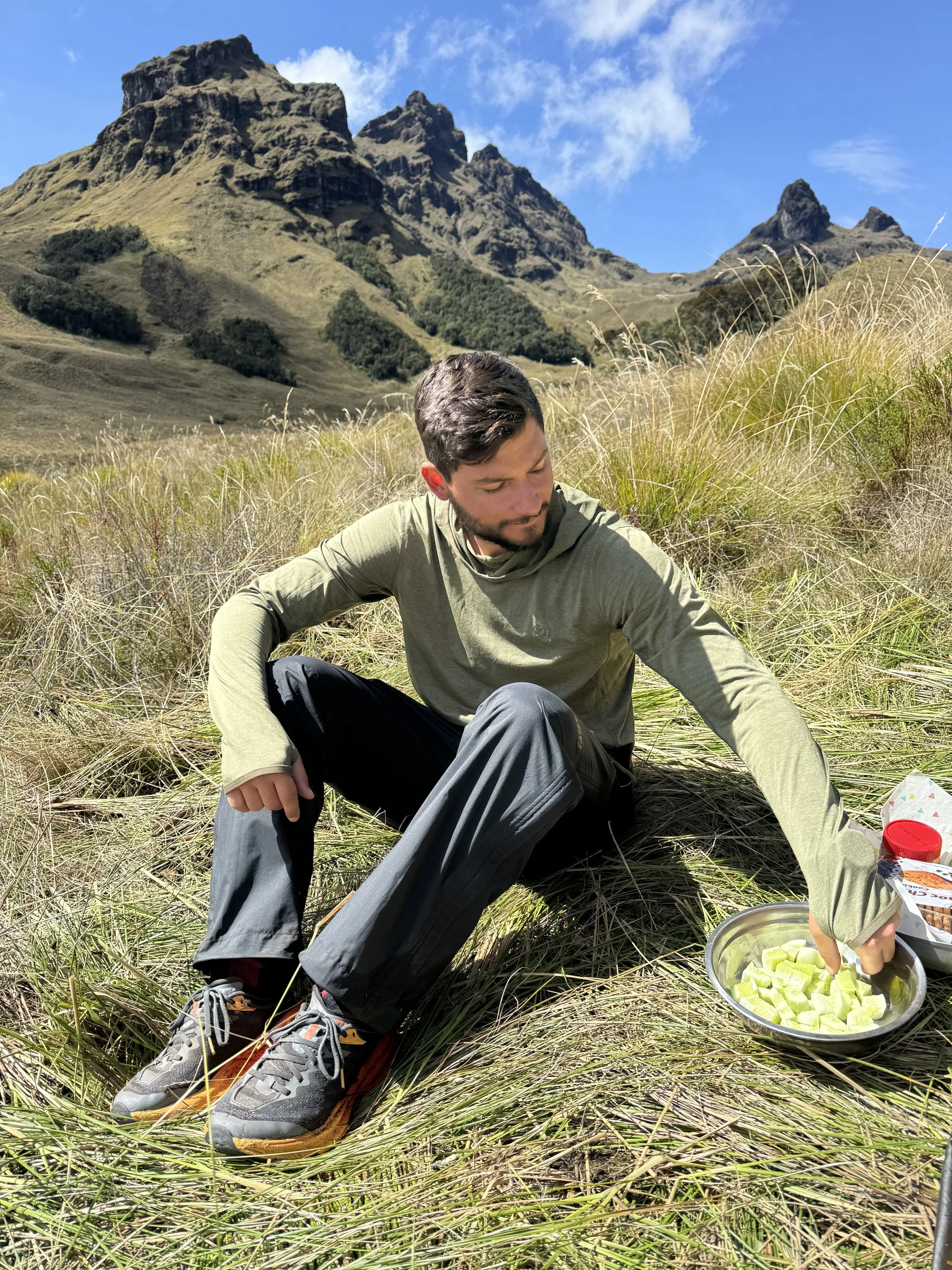 A man in outdoor clothing is sitting on the grass in a field with tall grass and mountains in the background under a blue sky. He is reaching into a bowl of sliced cucumber on the ground.