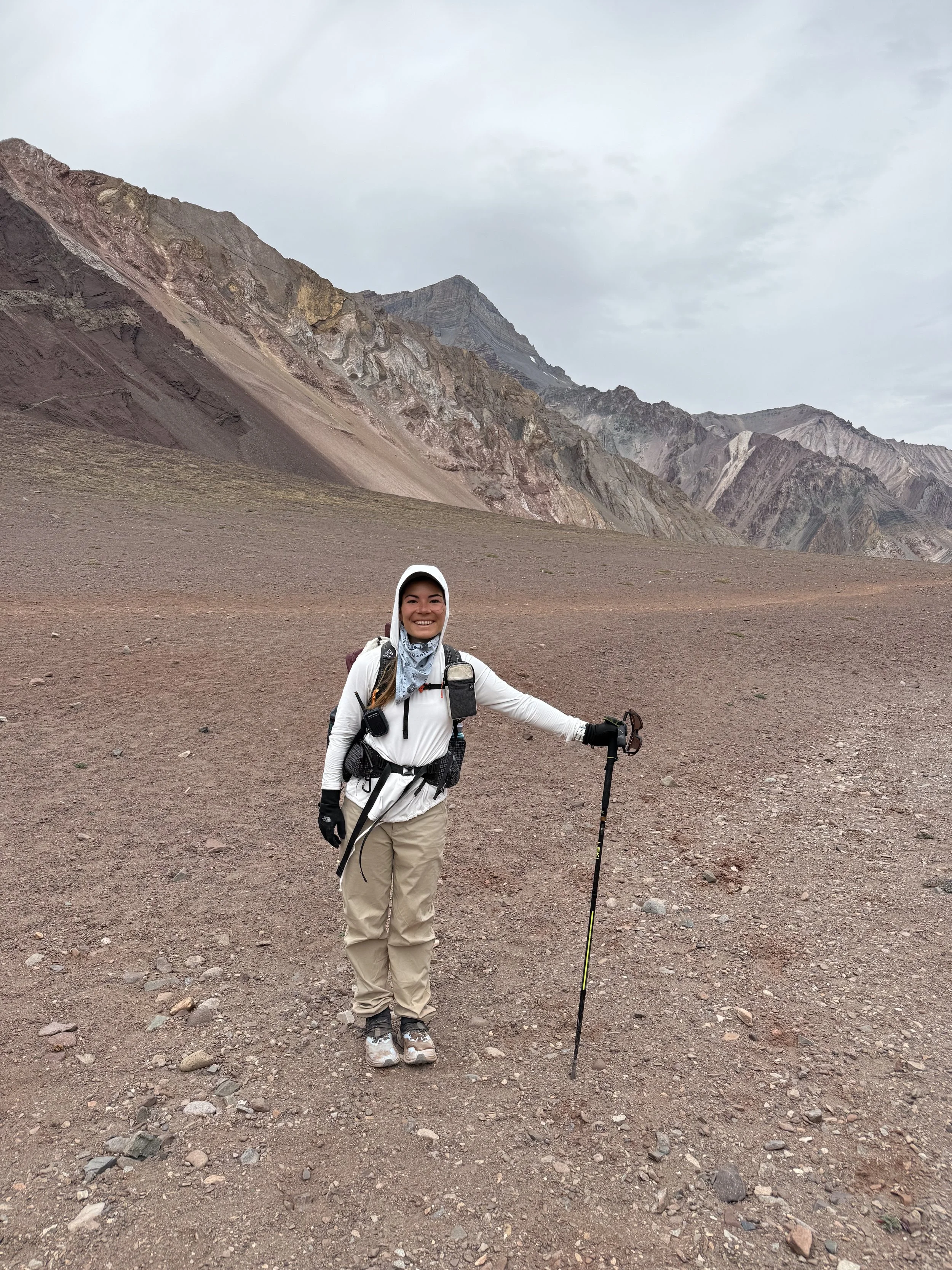 A woman dressed in outdoor hiking gear standing on a rocky dirt landscape with mountains in the background, holding a hiking stick and smiling.