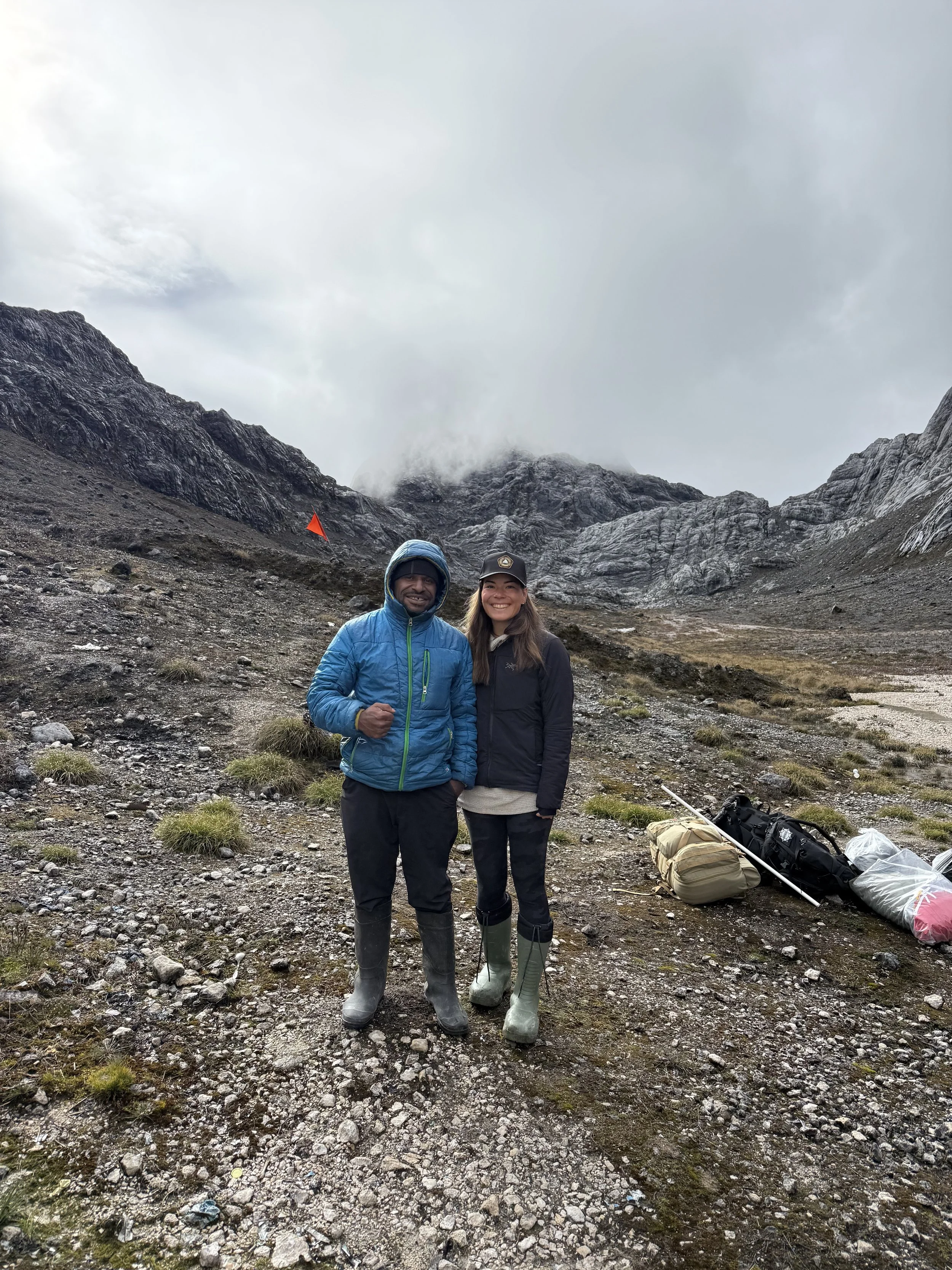 Two hikers dressed in outdoor gear standing on rocky terrain with mountains and cloudy sky in the background, carrying camping supplies.
