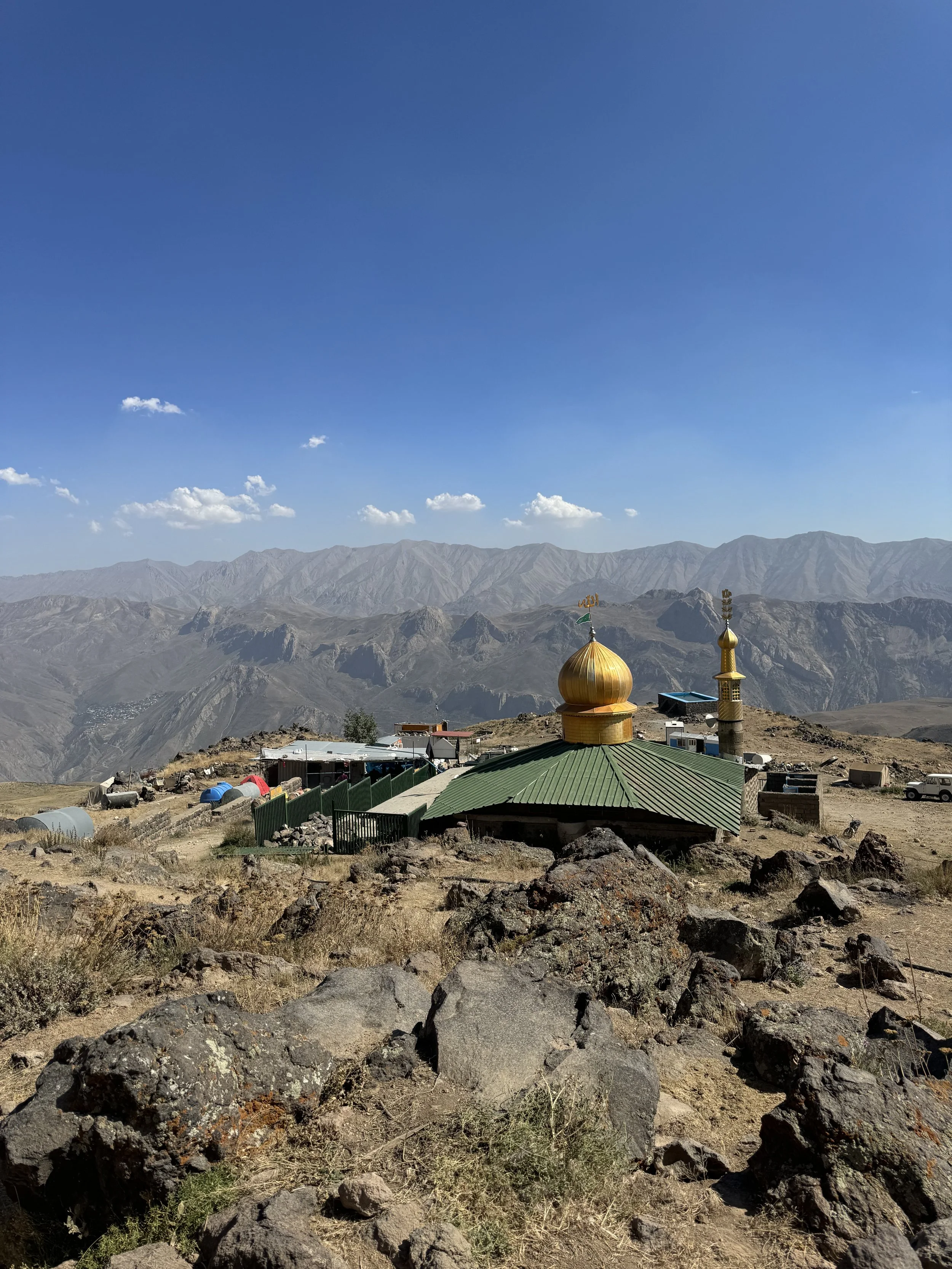 A small mosque with a golden dome and a minaret located on a rocky, mountainous landscape under a blue sky with scattered clouds.