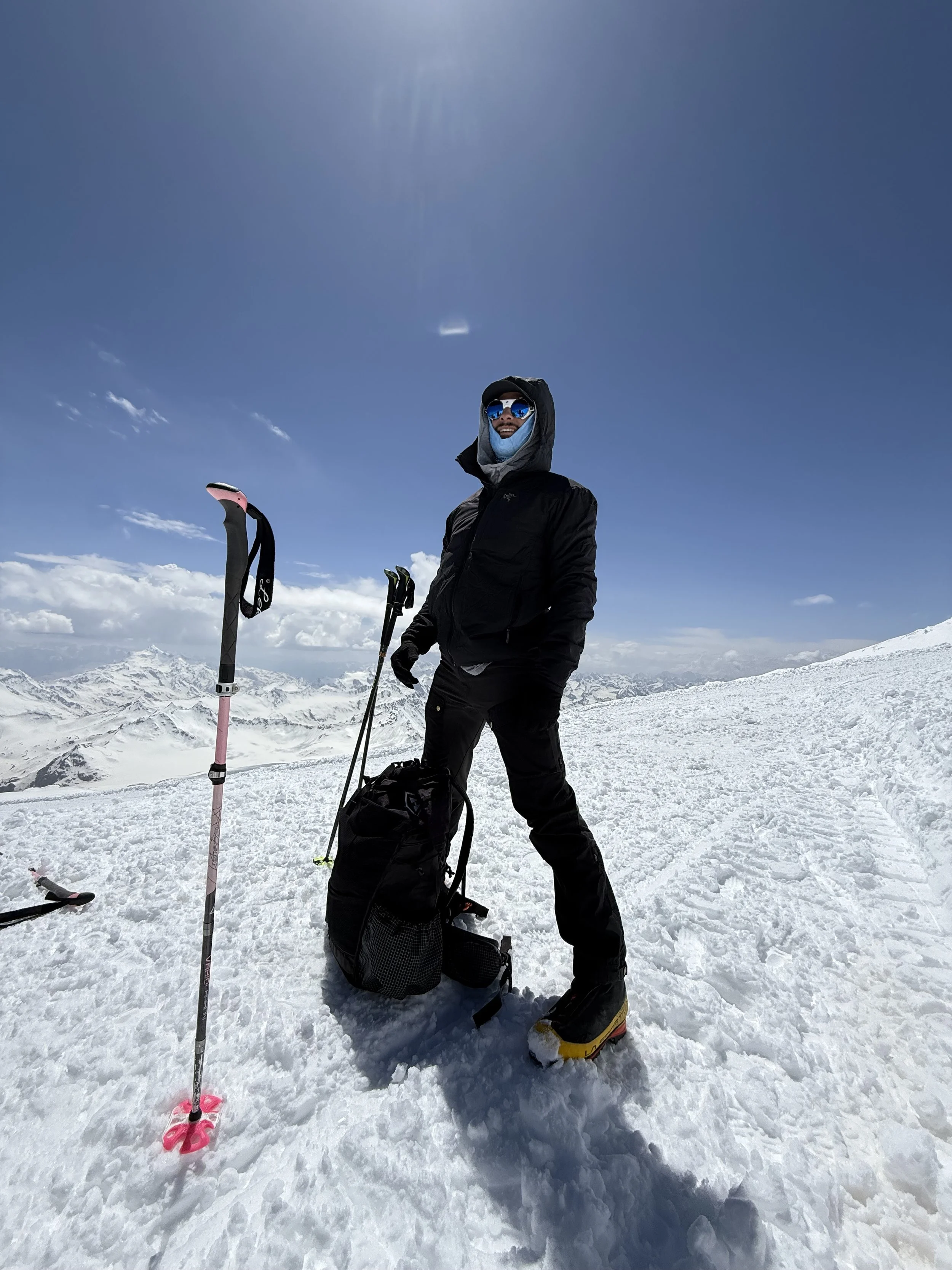A person dressed for snow hiking or mountaineering, standing on a snow-covered mountain with ski poles and a backpack, surrounded by snowy peaks under a clear blue sky.