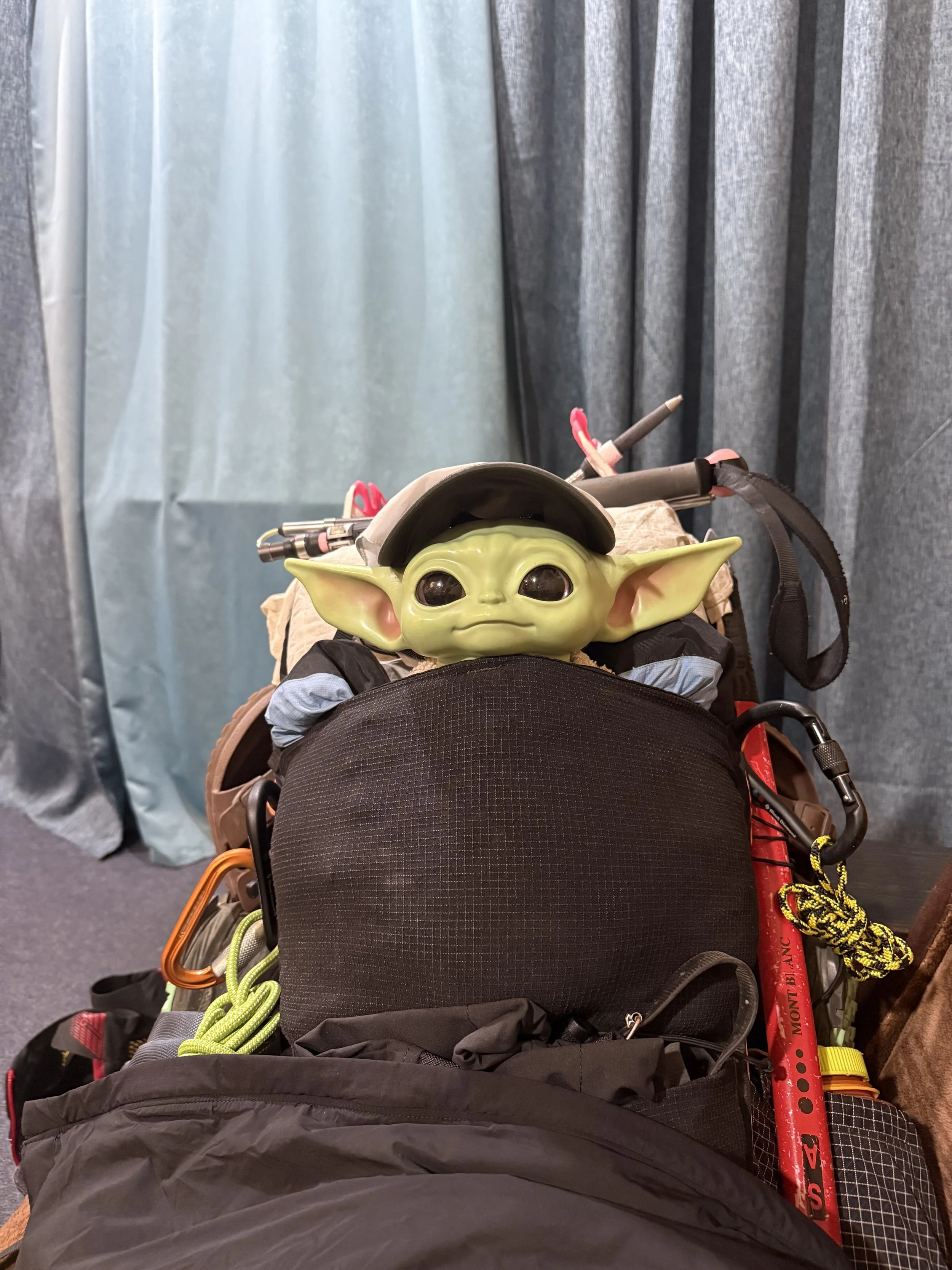 A Baby Yoda (Grogu) toy or figure peeking out from a backpack or bag, with dark curtains in the background.