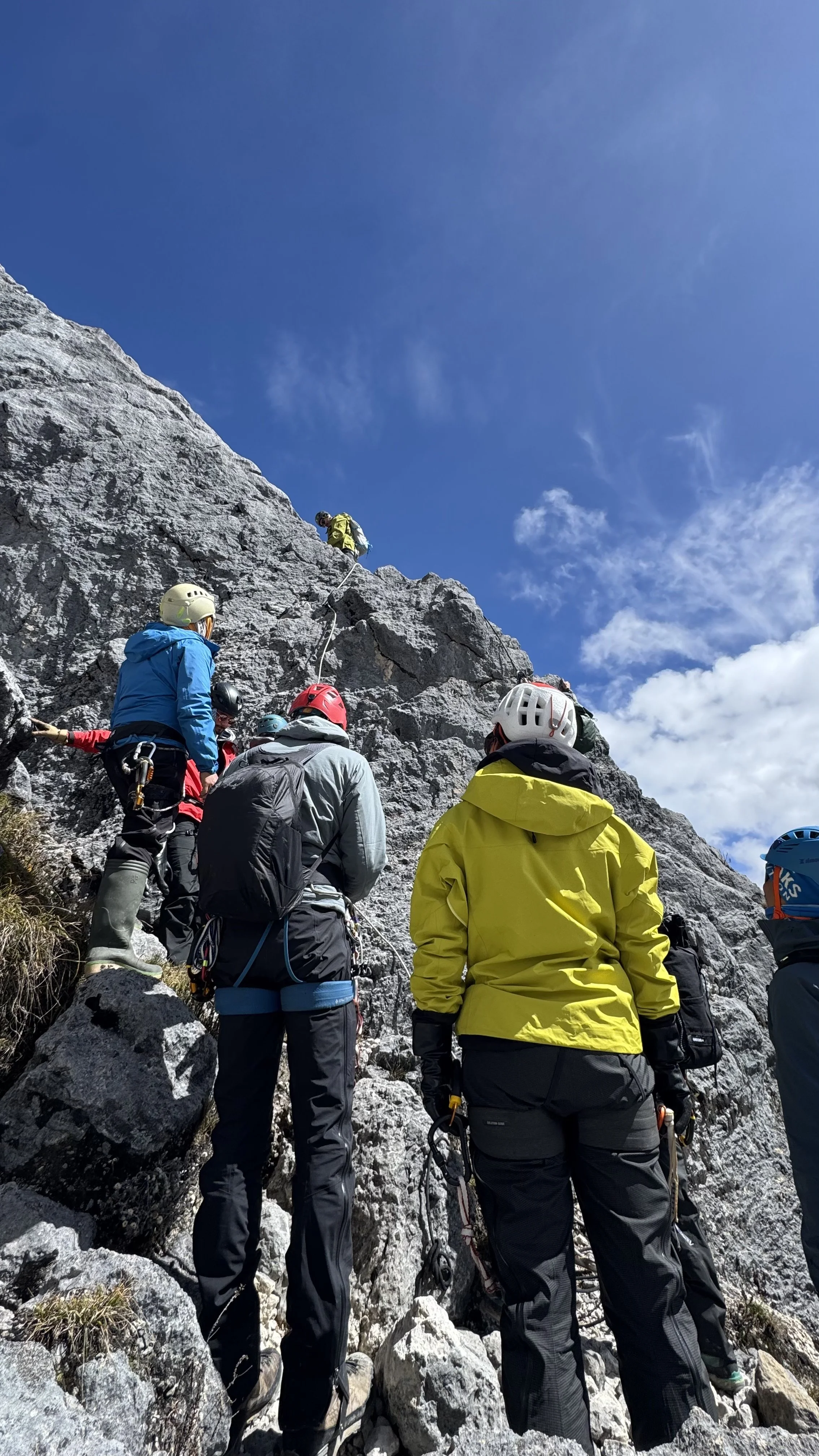 Group of climbers ascending a rocky mountain face, wearing helmets and outdoor gear, with one climber at the top securing a rope.