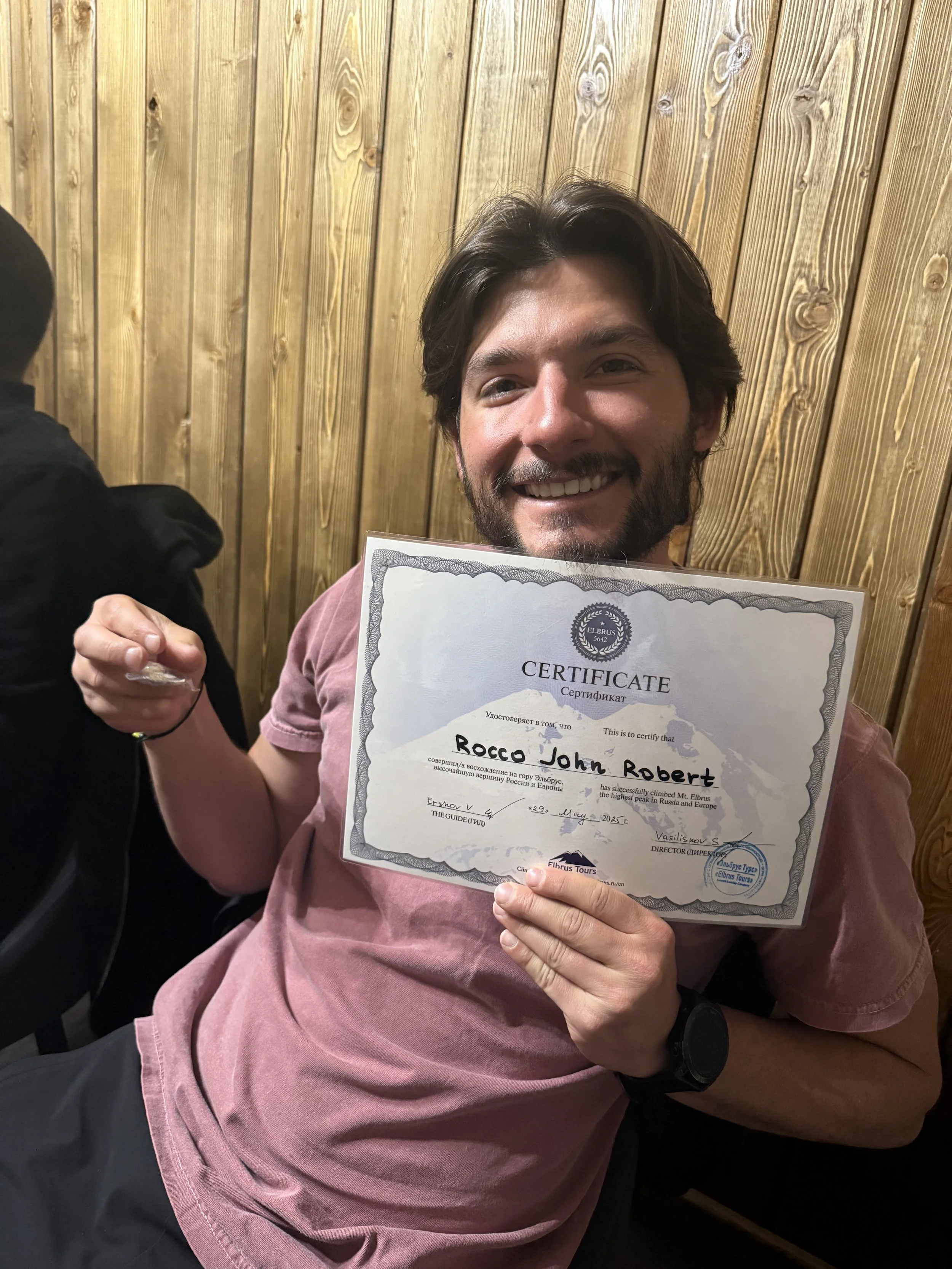 A smiling man with dark hair and beard holding a certificate in front of a wooden wall.