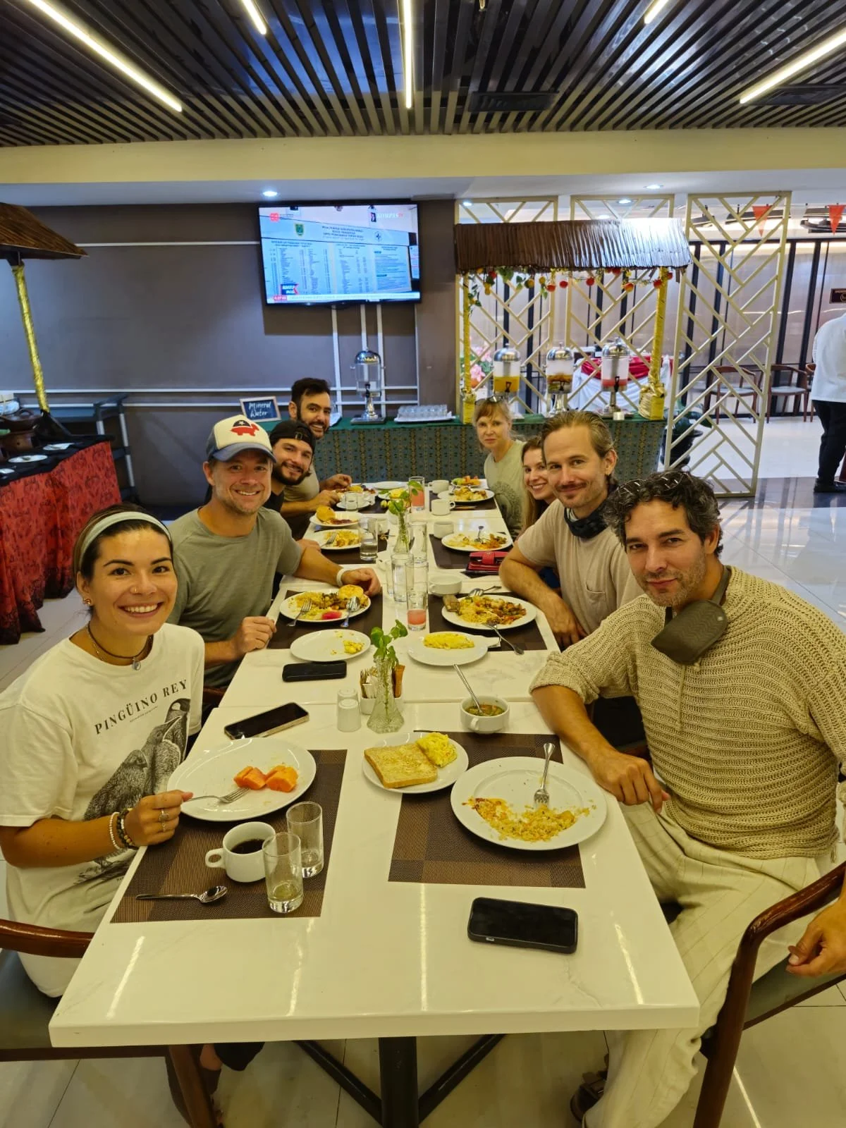 A group of nine people sitting around a dining table at a restaurant, with plates of food, cups, and glasses, smiling at the camera, in a well-lit indoor setting.