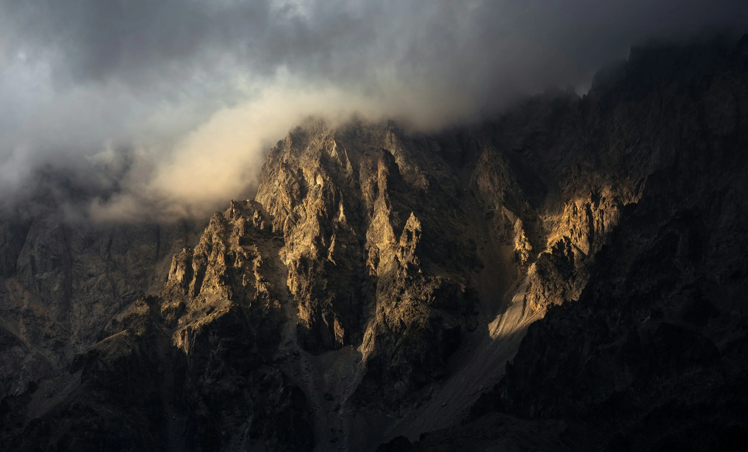 Dark mountain peaks partially shrouded in clouds with sunlight illuminating parts of the rocky surface.