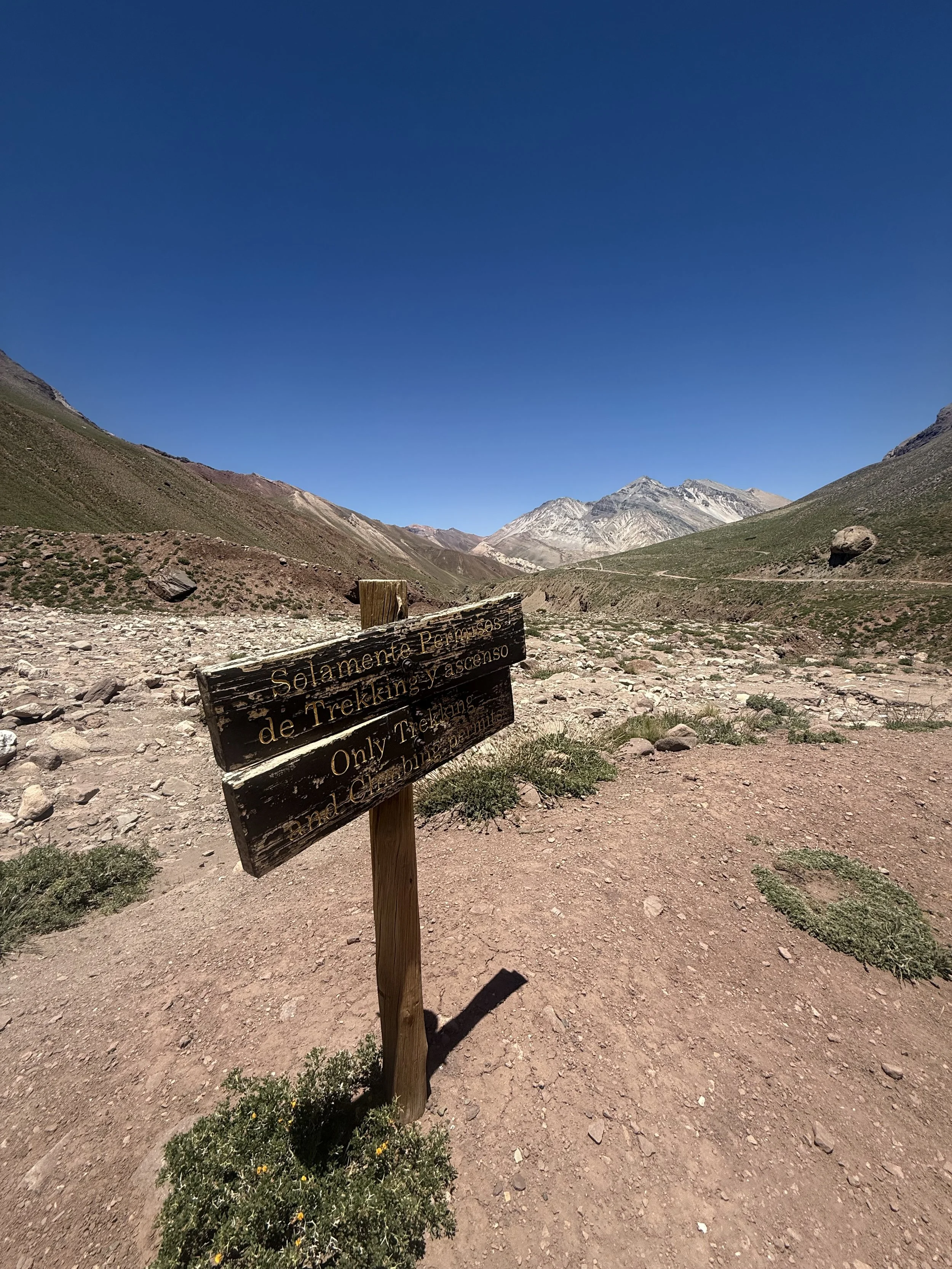 Mountain trail with a wooden sign that reads 'Solamente Permiso de Trekking y Ascenso' and 'Only Trekking and Climbing Permitted,' in a rocky, arid valley surrounded by mountains with a clear blue sky overhead.