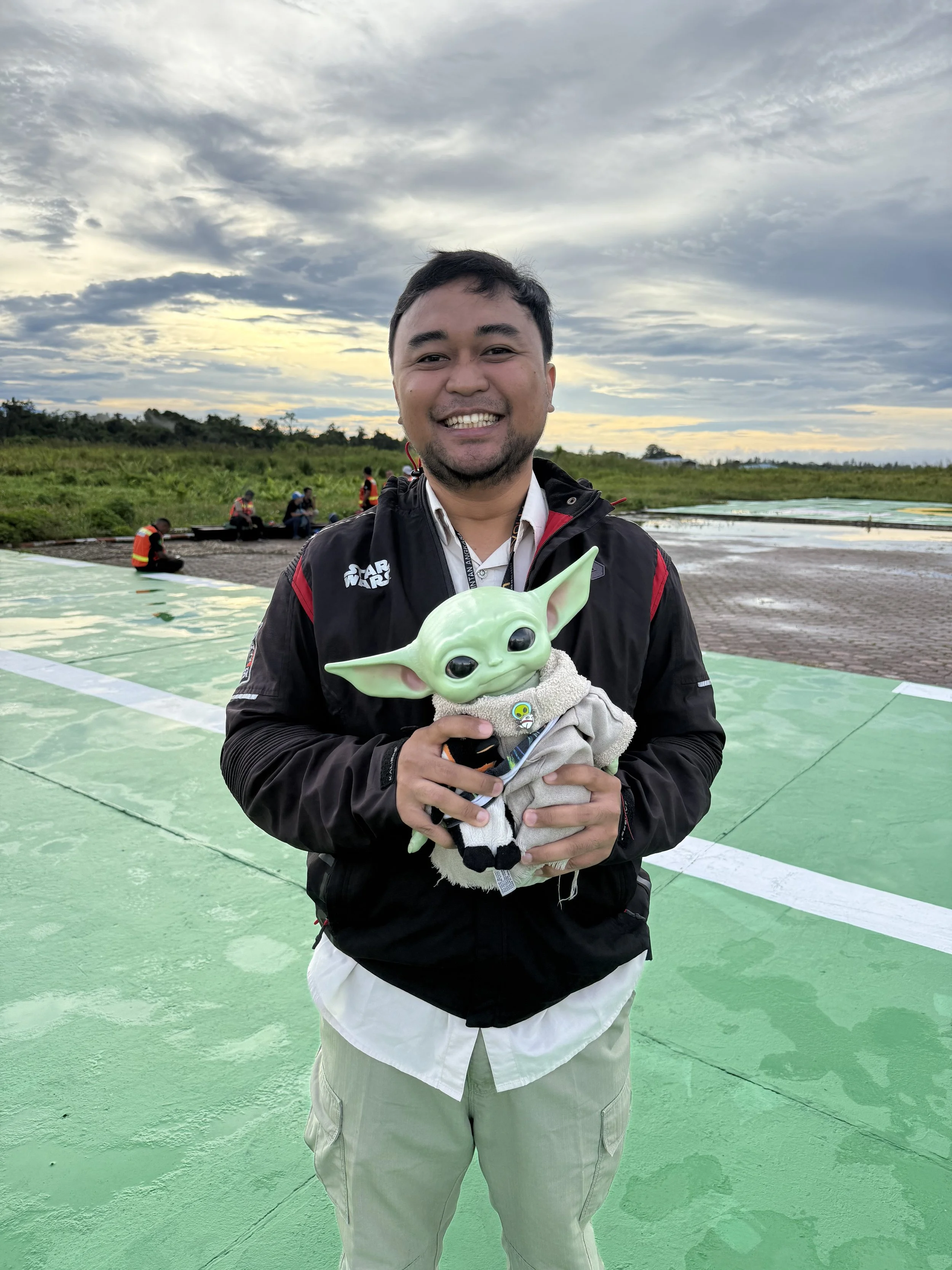 A man smiling outdoors on a cloudy day, holding a plush toy of Grogu (Baby Yoda) from Star Wars, standing on a green surface with other people in the background.