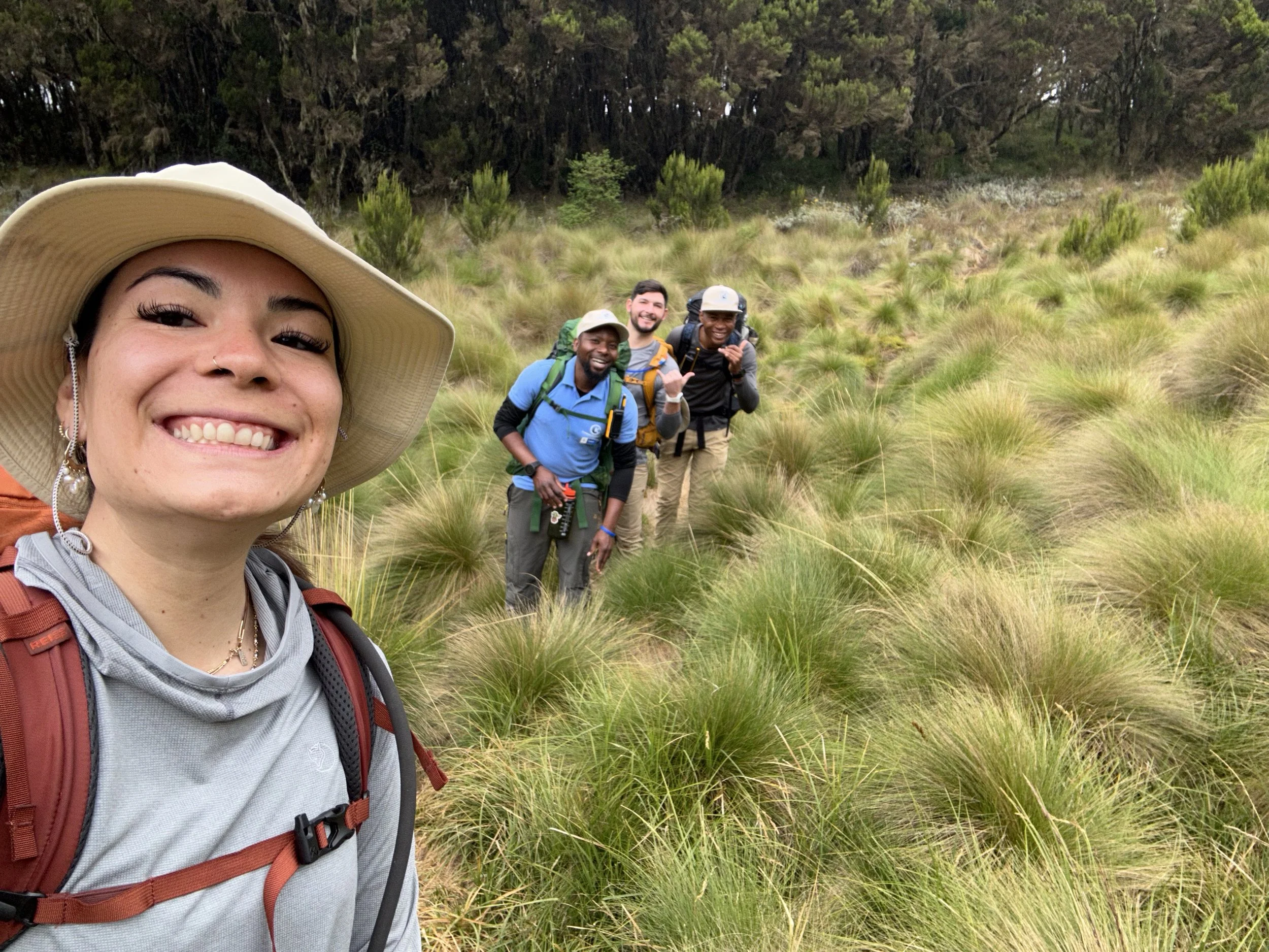 A woman wearing a wide-brimmed hat and a gray shirt taking a selfie with a group of four men behind her, hiking through a grassy outdoor area with bushes and trees in the background.