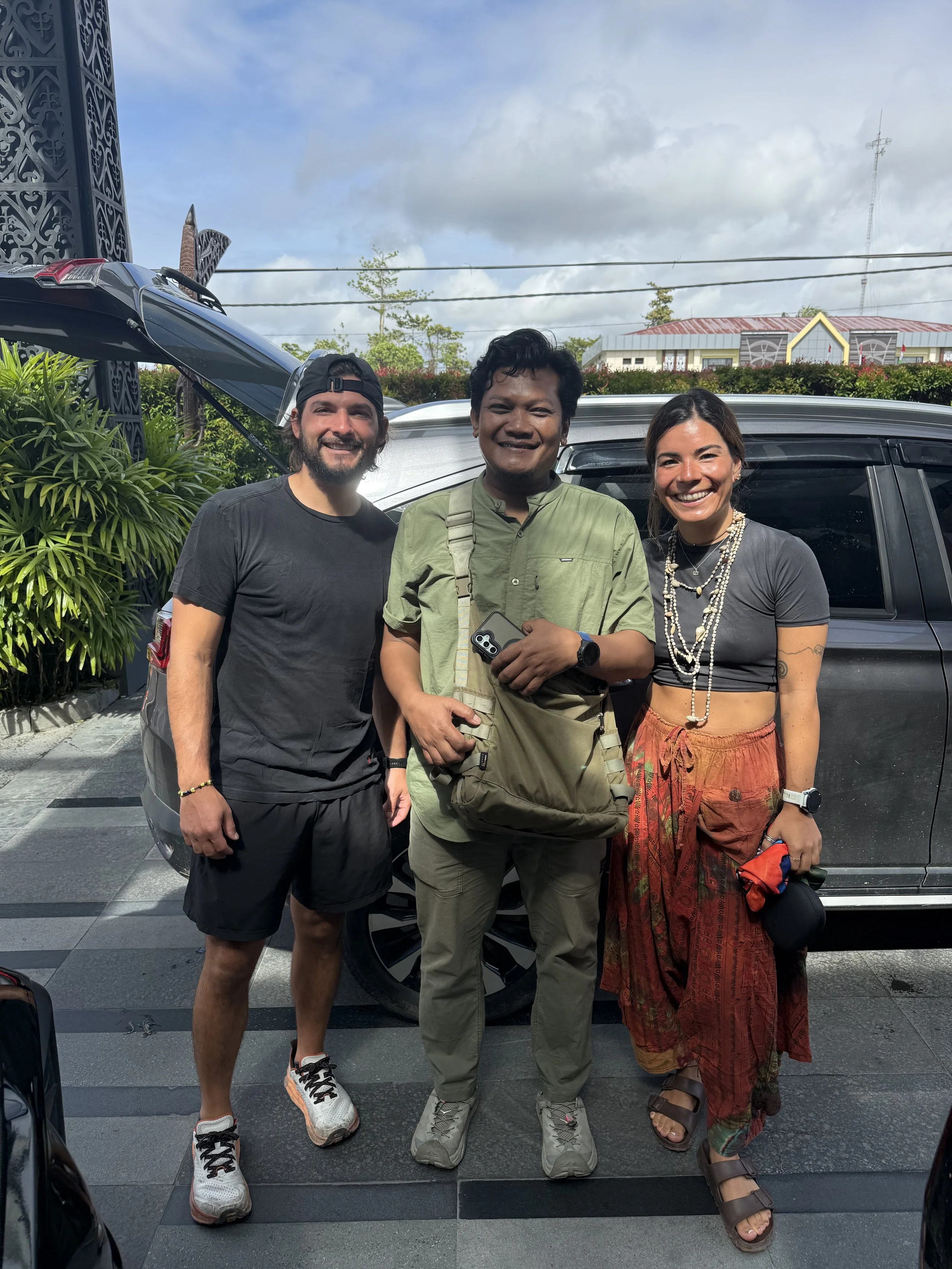 Three people standing together outdoors in front of a vehicle, smiling at the camera, with a cloudy sky and some greenery in the background.