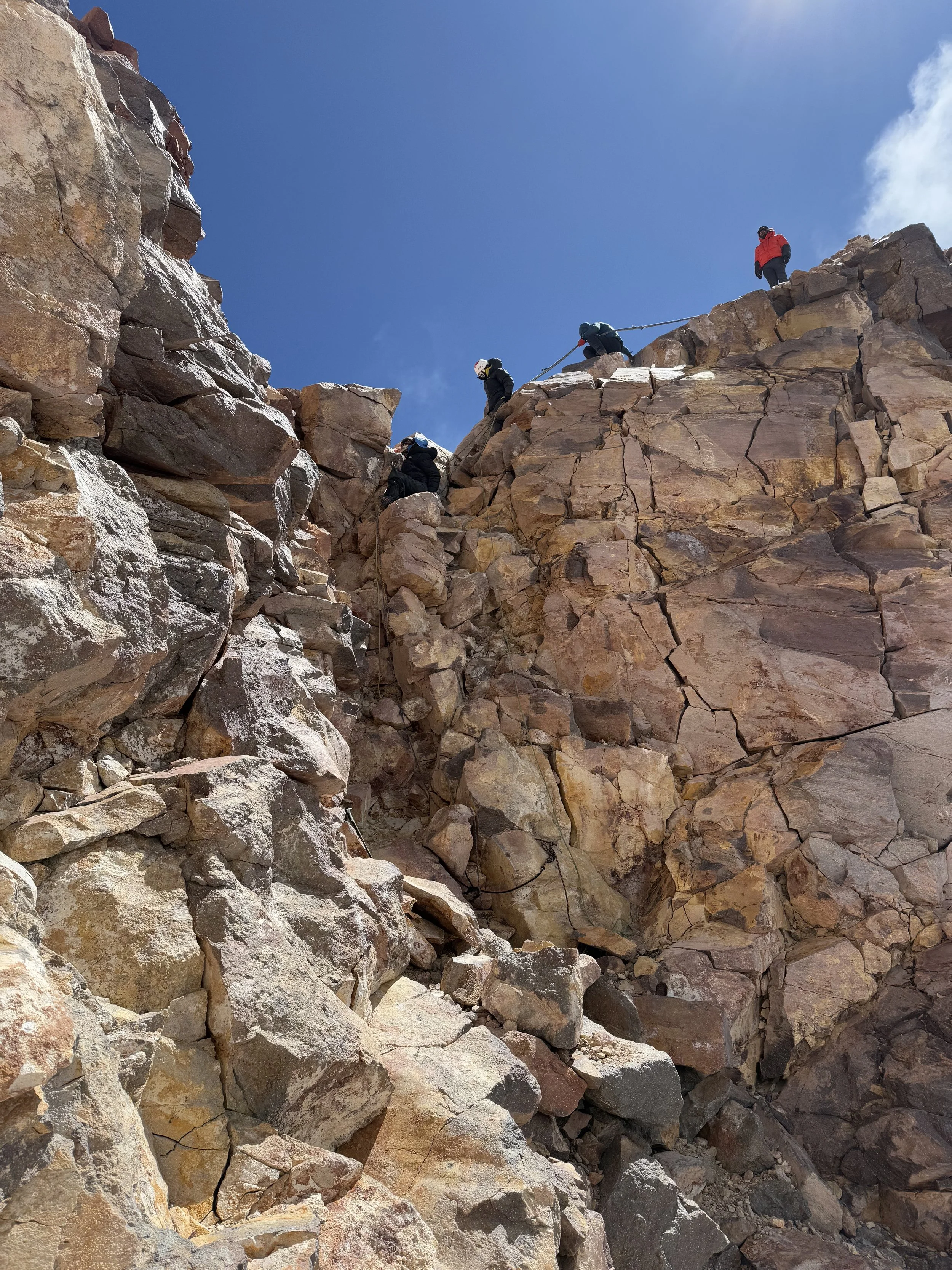 People rock climbing up a steep, rocky mountain under a clear blue sky.