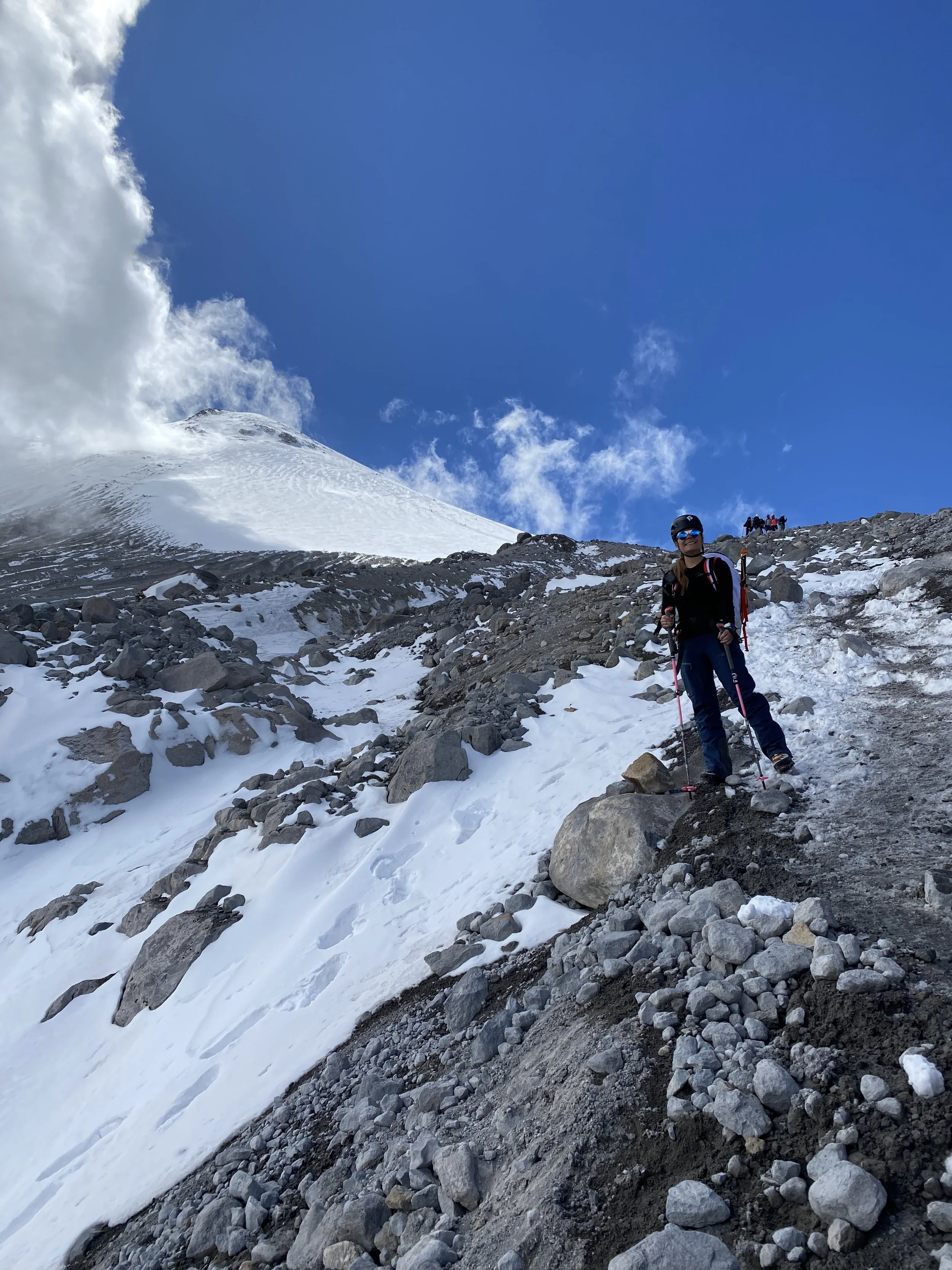 A person hiking on a snowy mountain trail with a blue sky, clouds, and a snow-covered volcano in the background.