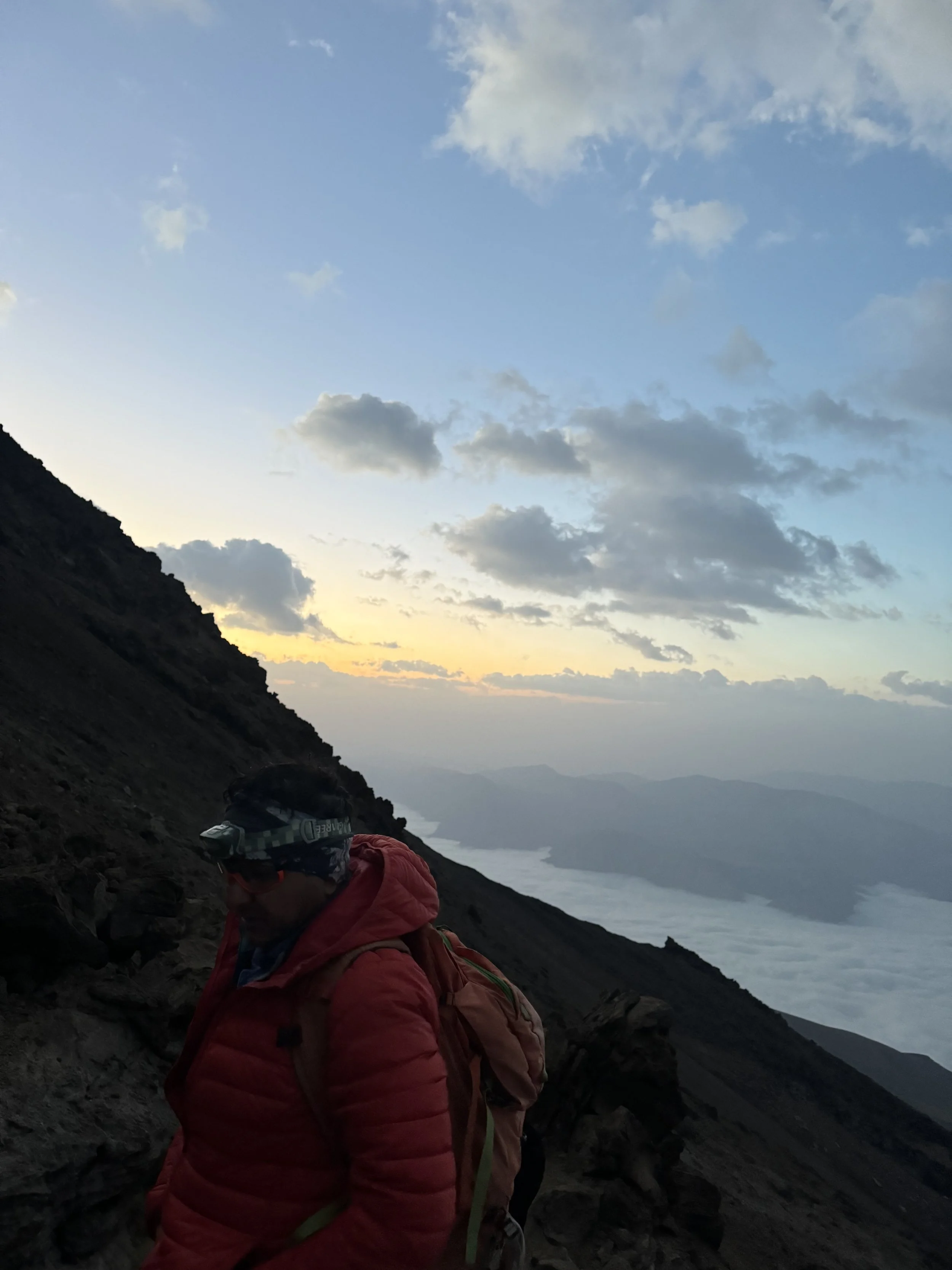 A person wearing a red jacket, black headlamp, and carrying a backpack, hiking on a mountain trail during sunset, with a view of clouds and distant mountains in the background.