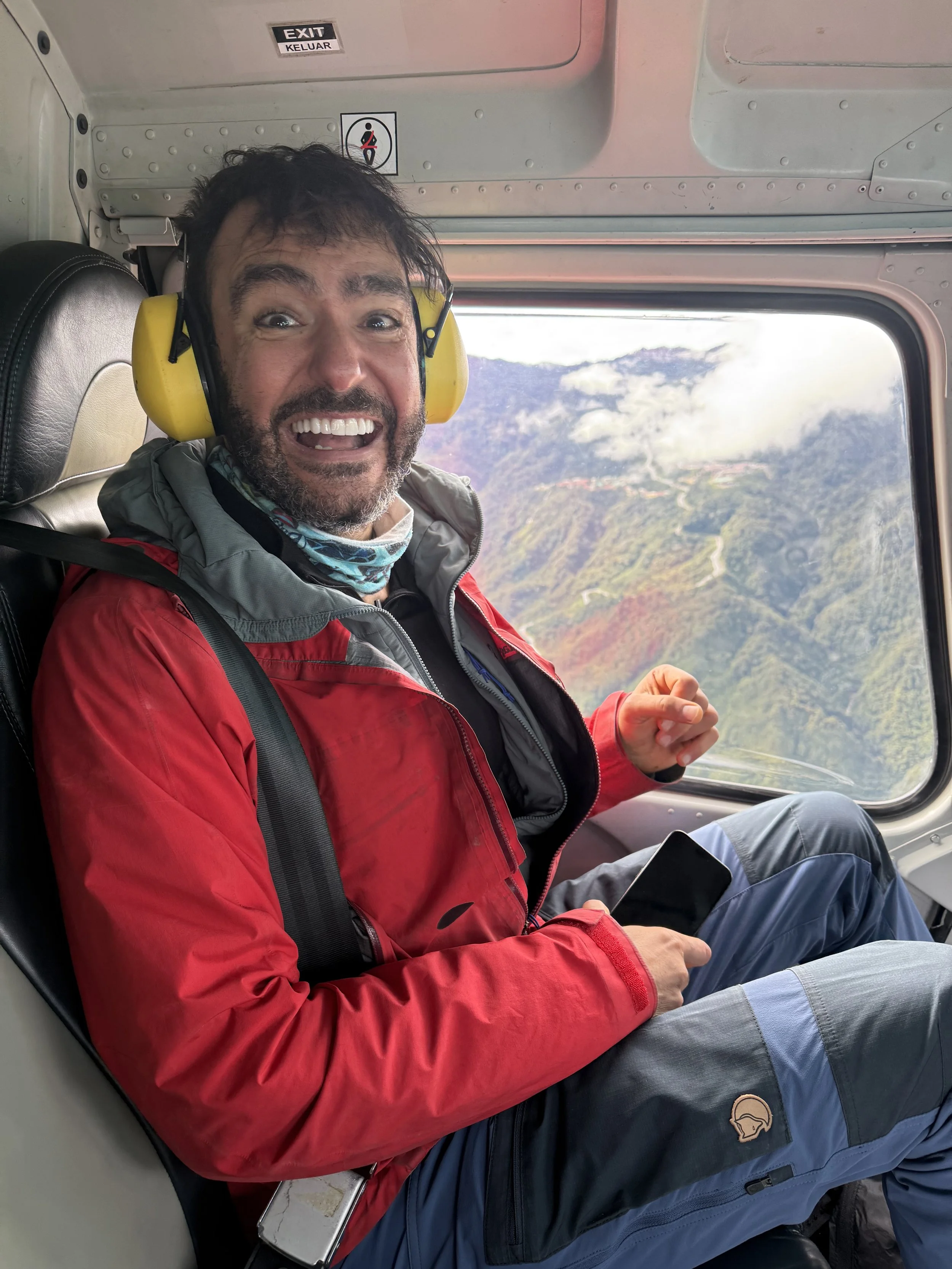 A man with a beard and short dark hair wearing a red jacket and yellow noise-canceling headphones, sitting inside a helicopter with a mountain landscape visible through the window behind him. He is smiling and holding a smartphone in his lap.