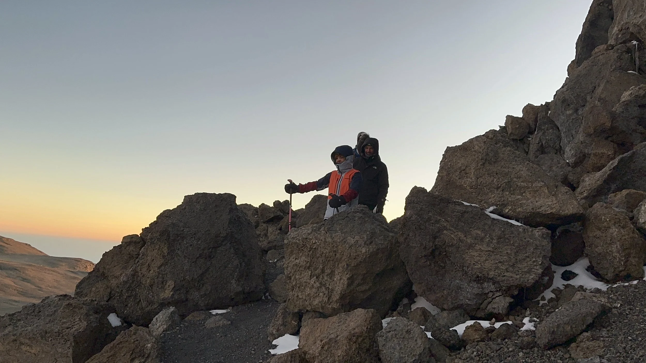 Two hikers in winter gear standing among large rocks on a mountainous terrain during sunset, with snow patches on the ground.