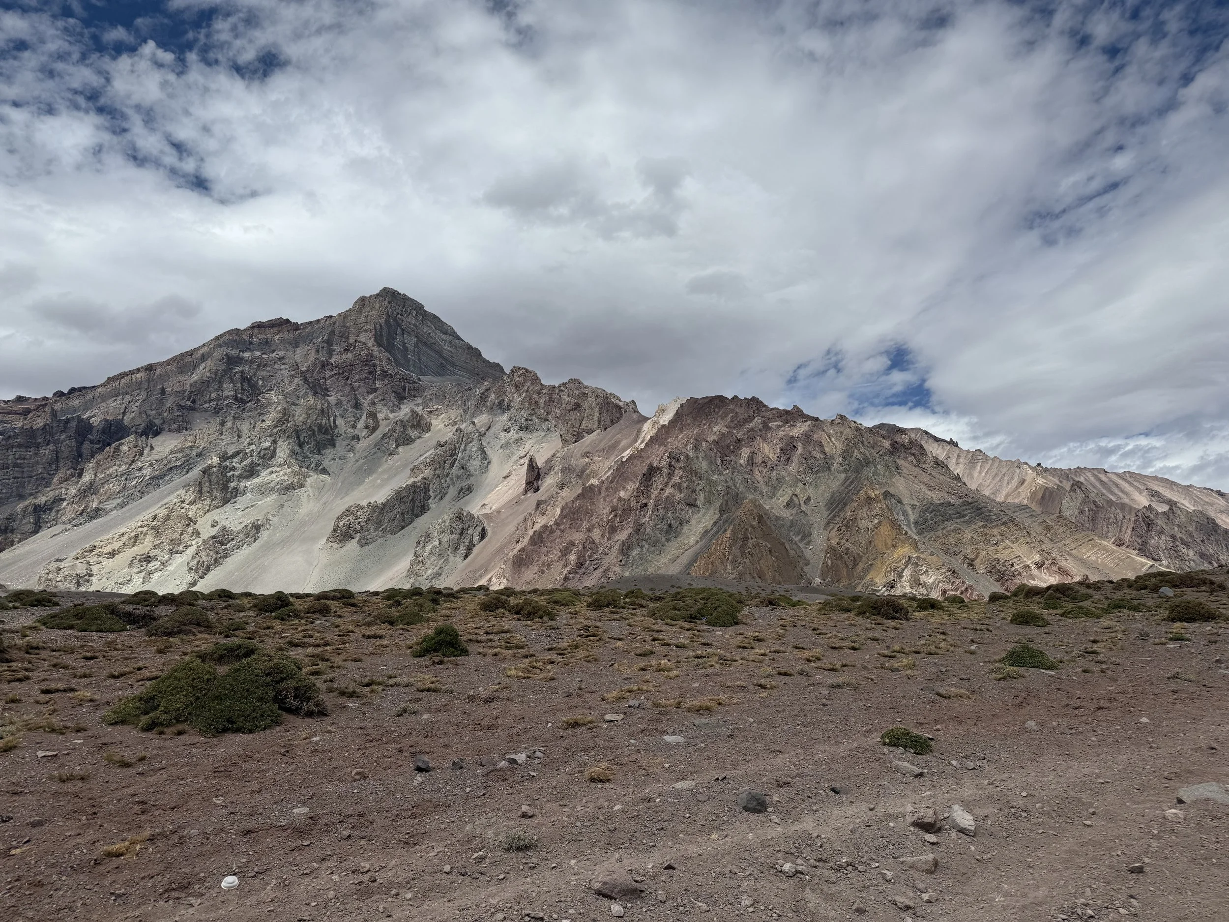 A dry, rocky landscape with sparse green shrubs in front of large, colorful mountain formations under a partly cloudy sky.