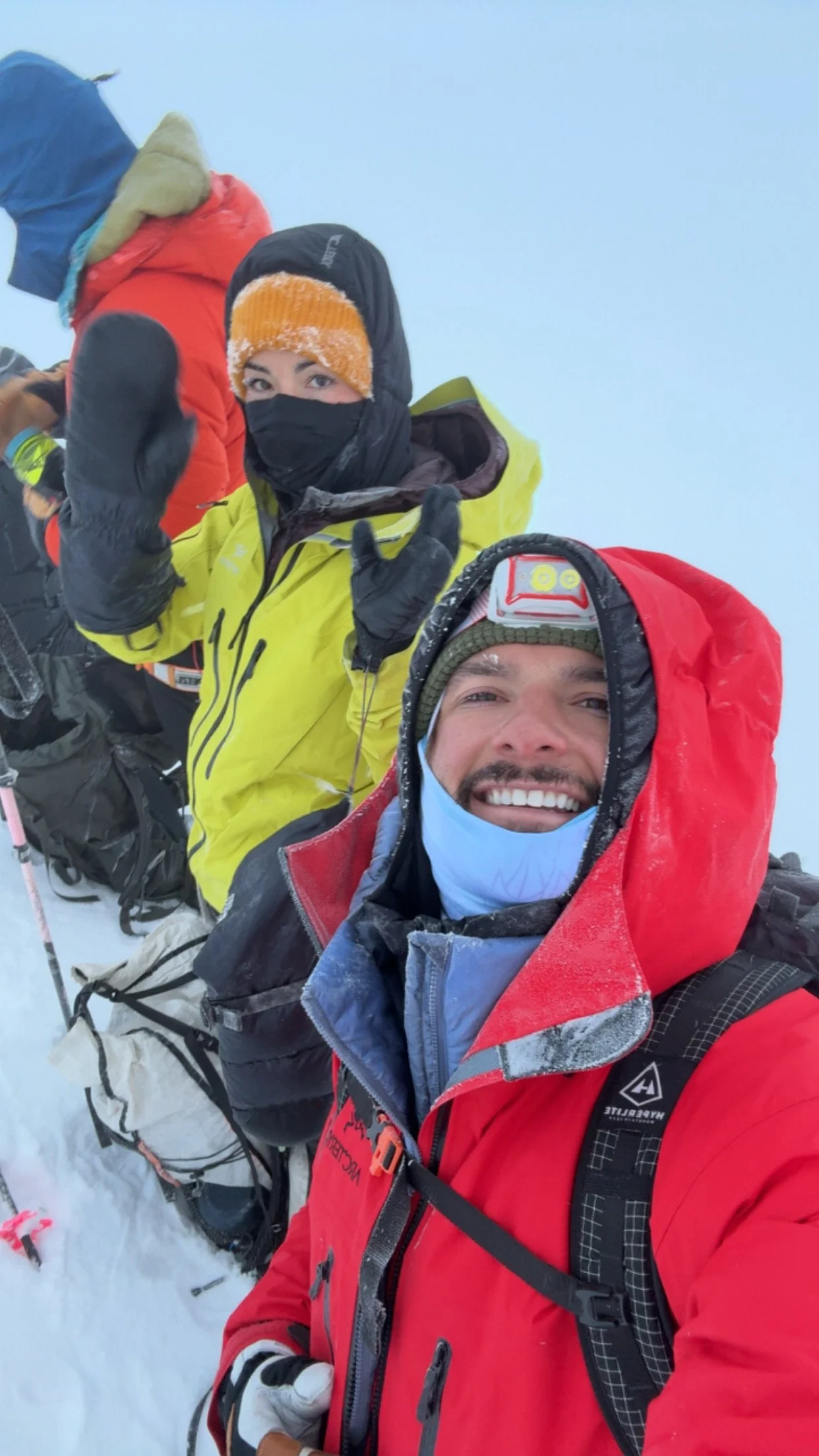 Two mountaineers in cold-weather gear and jackets, smiling and taking a selfie on a snowy mountain summit, with other climbers behind them.