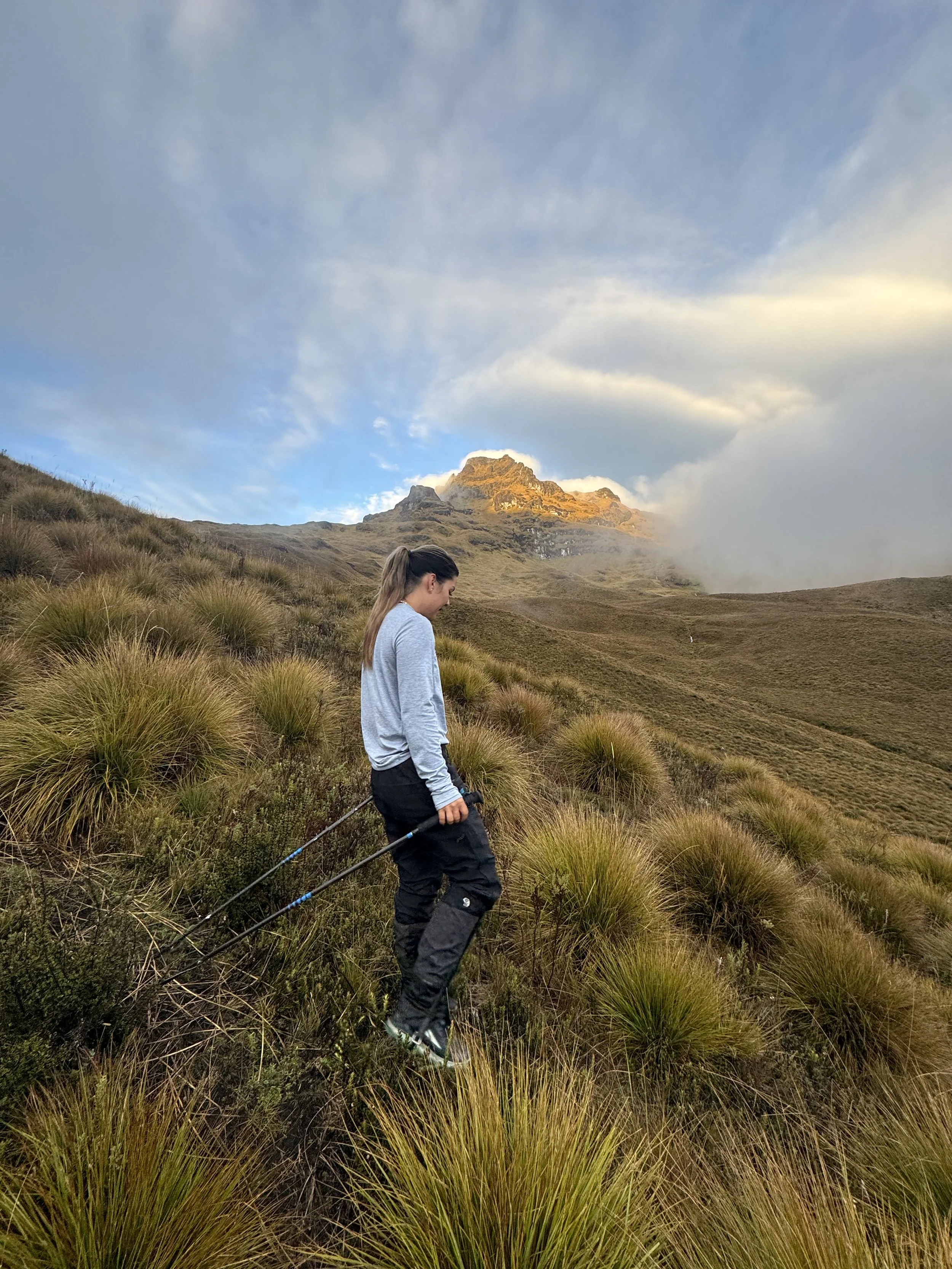 A woman hiking on a grassy hillside with mountainous terrain and a partly cloudy sky in the background.