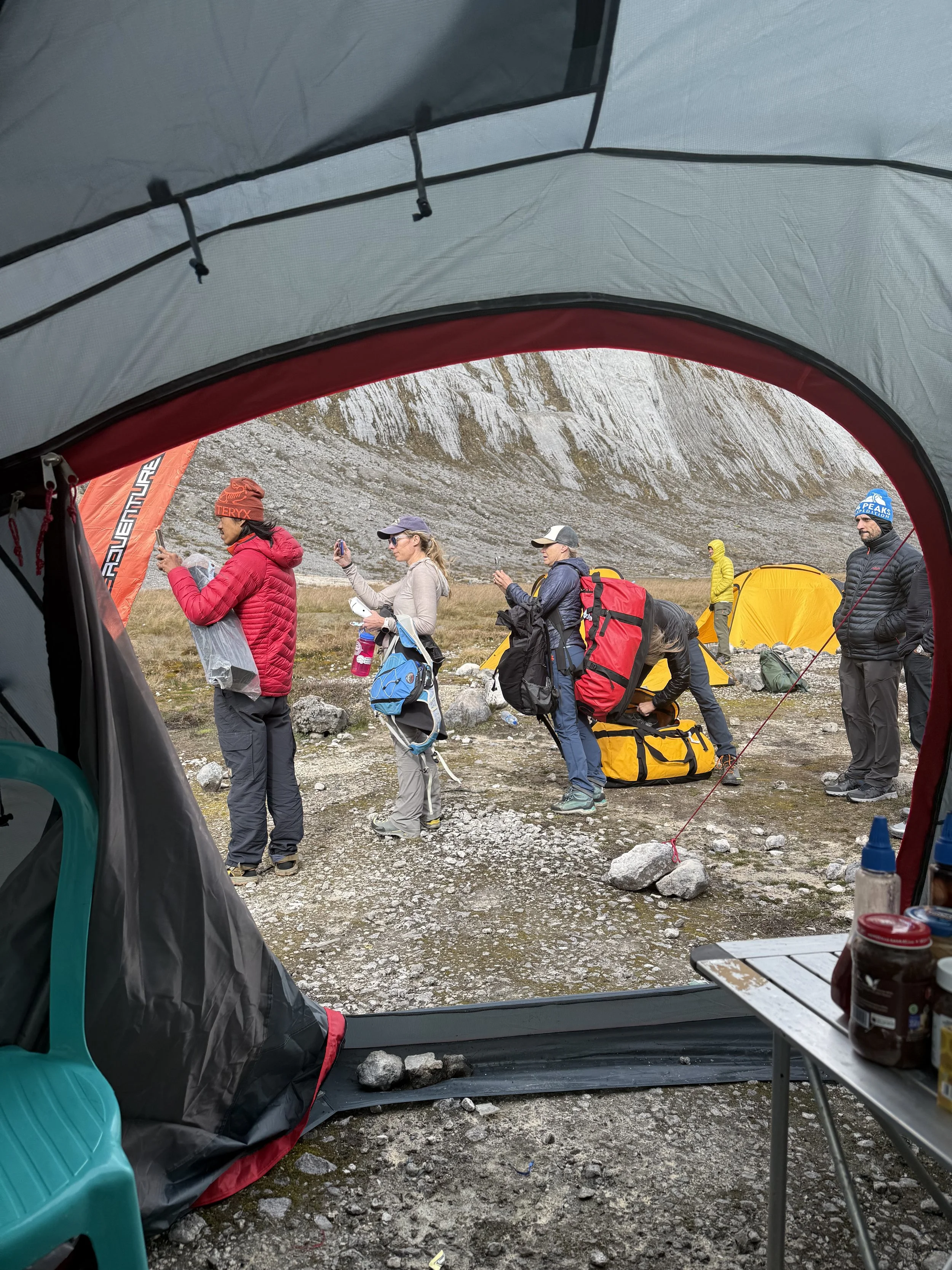 View from inside a tent looking out at a group of hikers preparing at a mountain campsite with tents on rocky terrain and a steep hillside in the background.