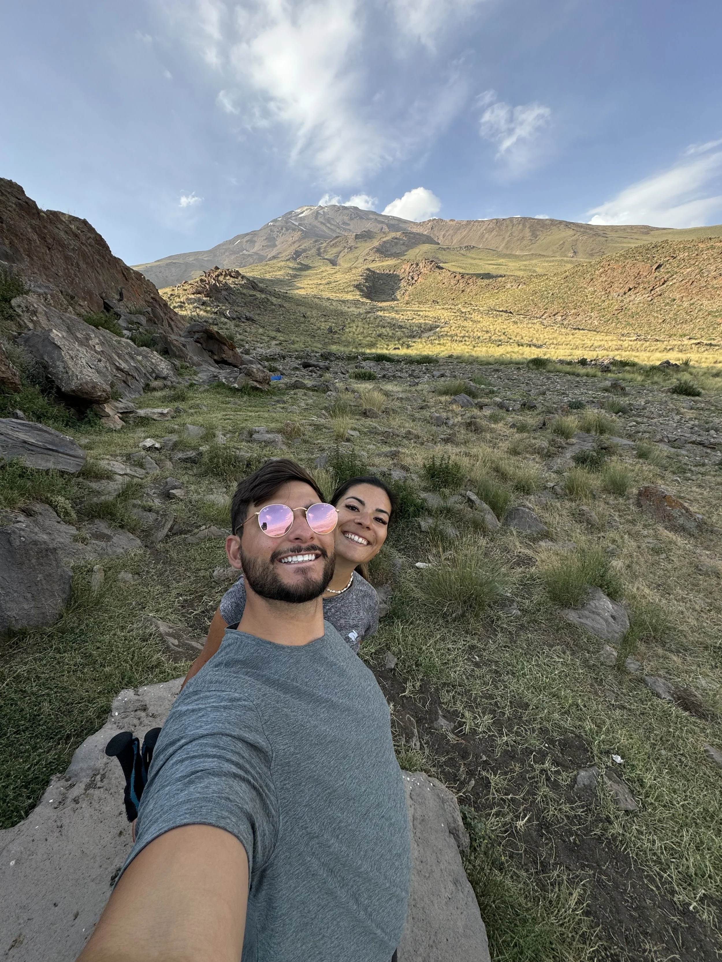 Two people smiling and taking a selfie in a mountainous outdoor landscape with grassy terrain and rocks, under a partly cloudy blue sky.
