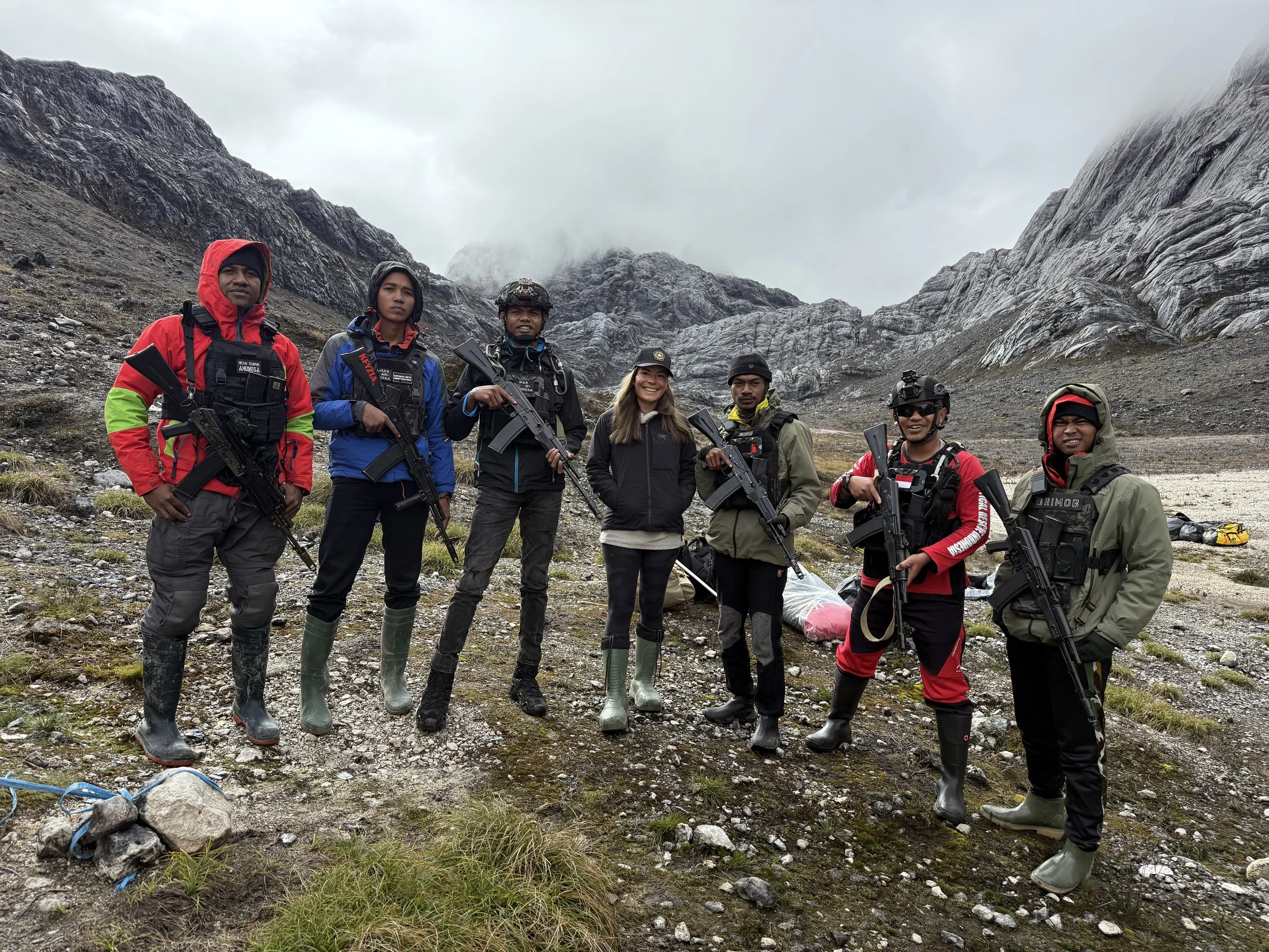 A group of seven people standing outdoors in a mountainous area with cloudy skies, wearing outdoor gear and holding rifles, smiling at the camera.