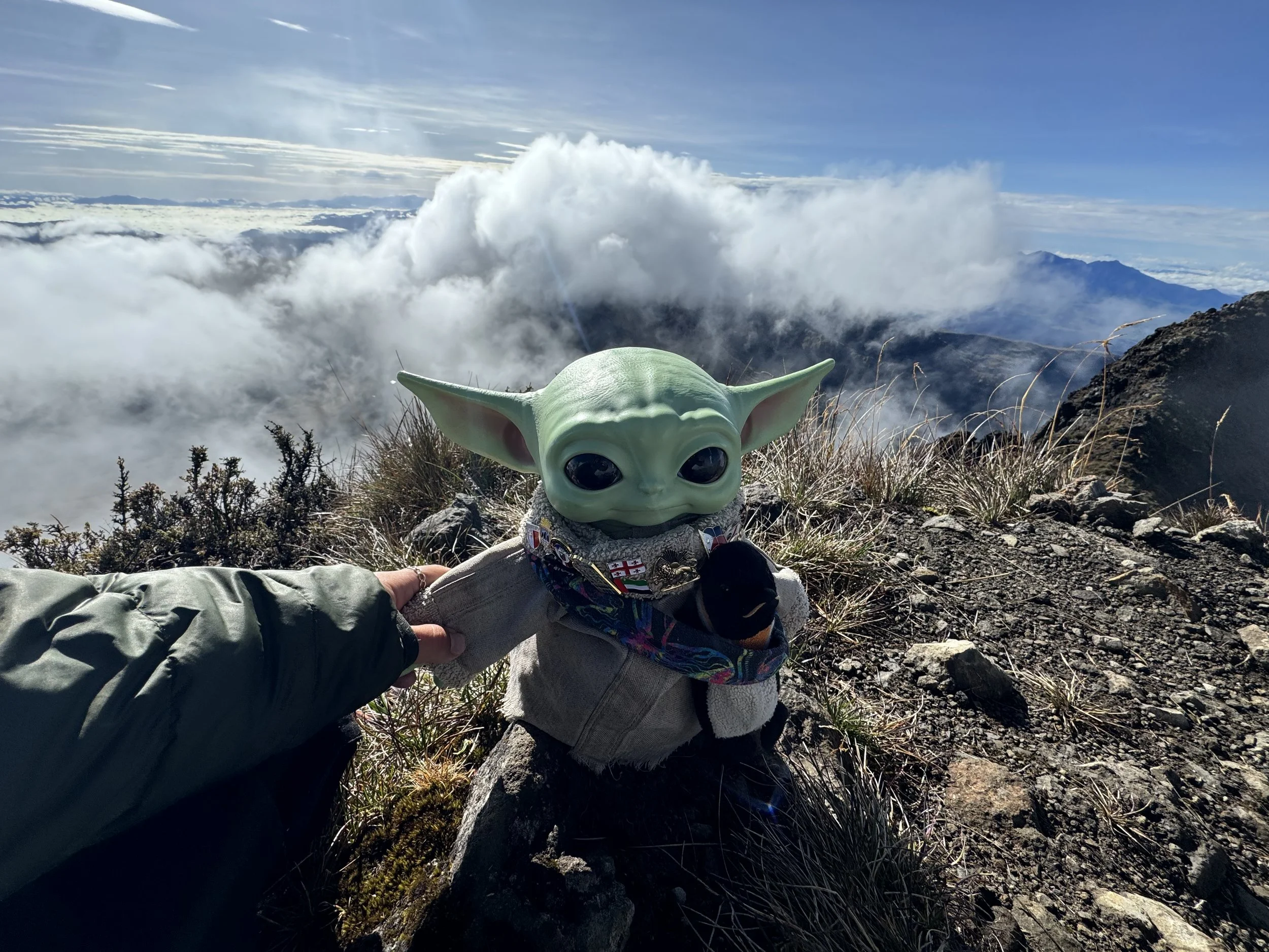 A person holding a plush toy of Baby Yoda at the top of a mountain with clouds and distant mountains in the background.