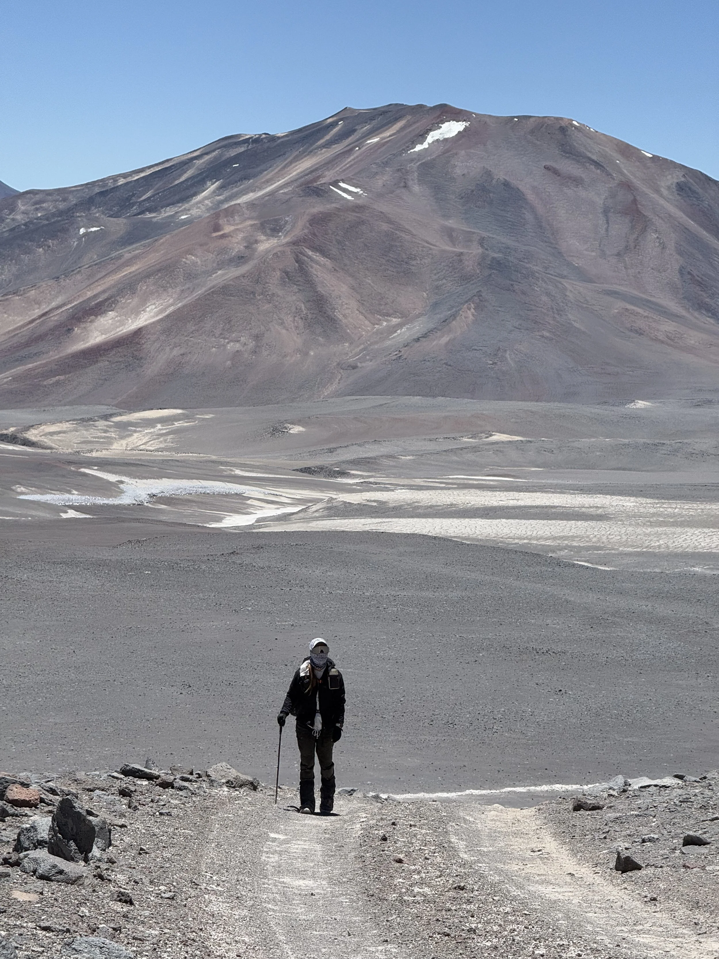 A hiker walking on a rocky trail in a barren, desert-like landscape with a large mountain in the background under a clear blue sky.