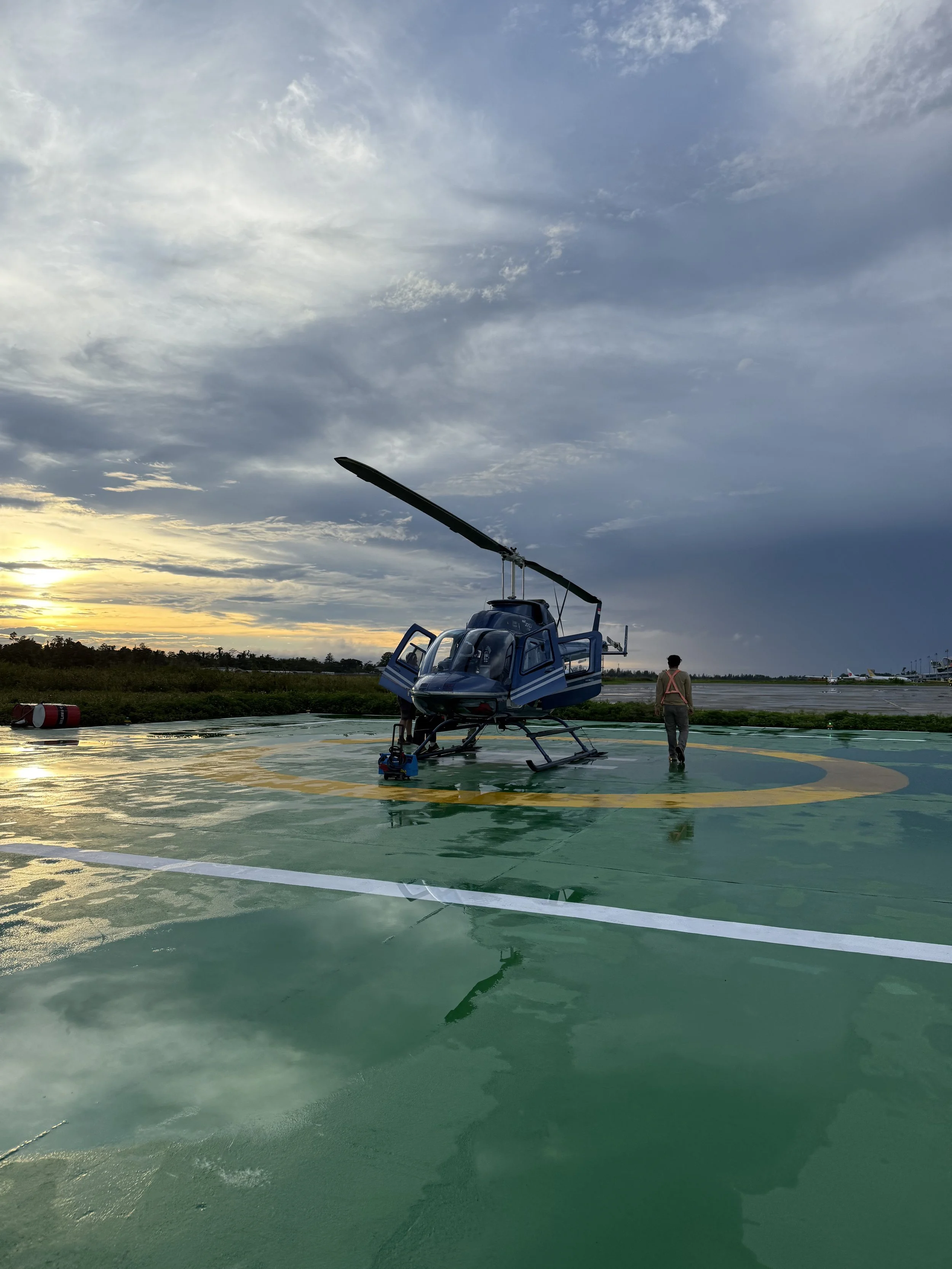 A helicopter parked on a helipad during sunset, with a person walking nearby and airport structures in the background.