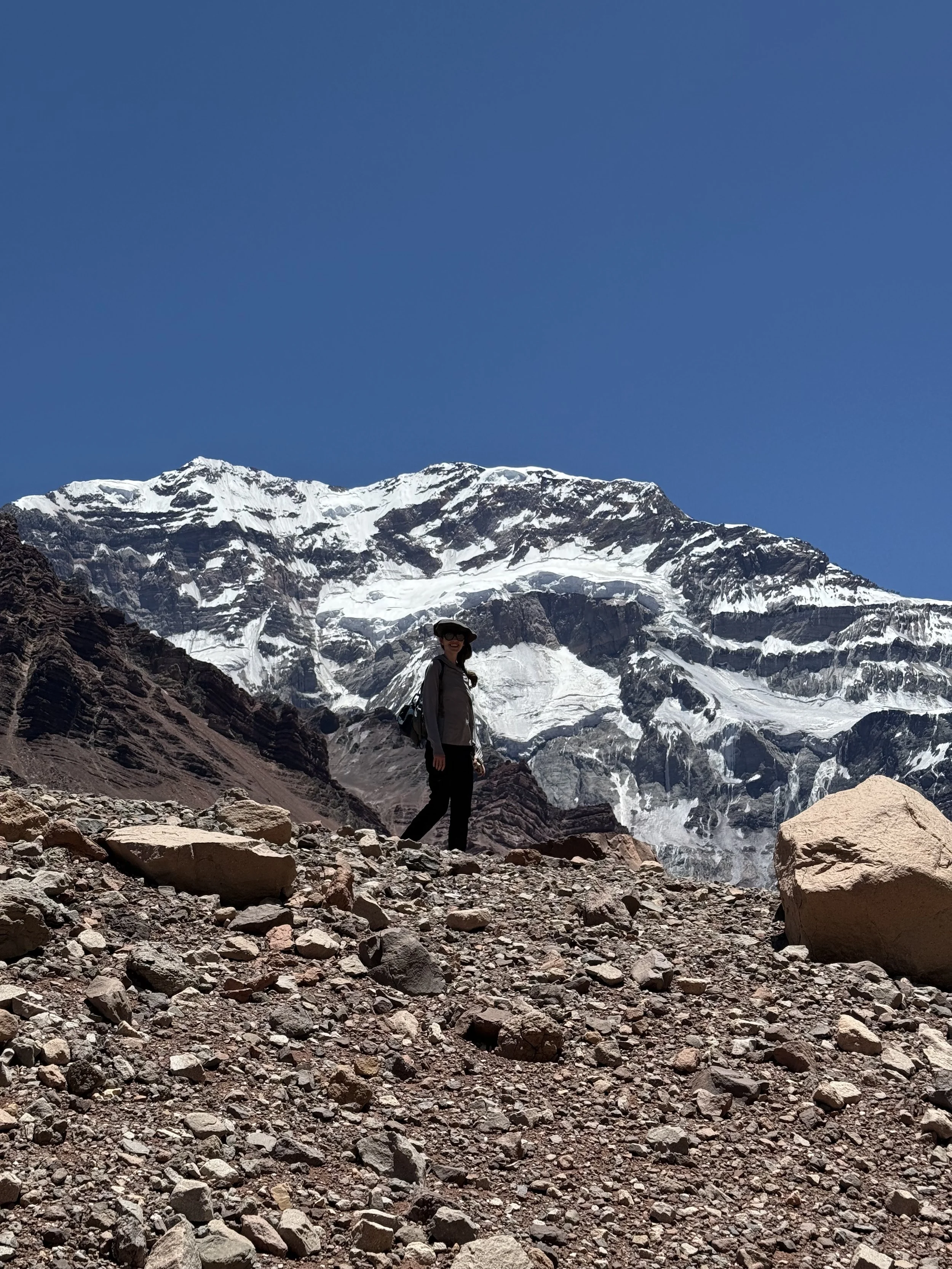 A woman standing on rocky ground in front of a snow-capped mountain with a clear blue sky.