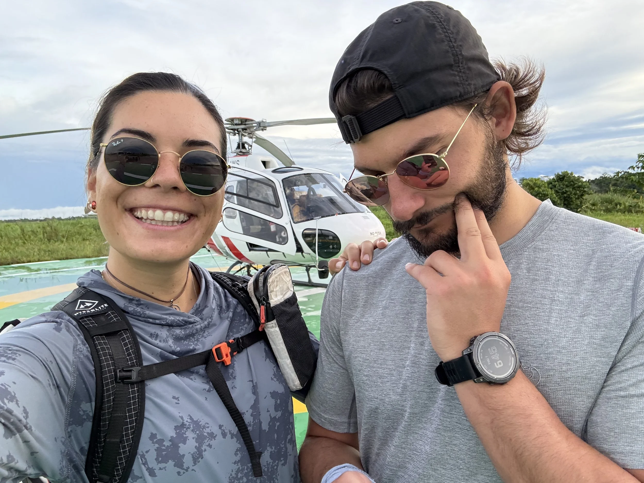 A woman with sunglasses and a man in a gray shirt and sunglasses stand in front of a helicopter in an open field. The woman is smiling, the man is looking down with a hand on his chin, and the helicopter is visible in the background.
