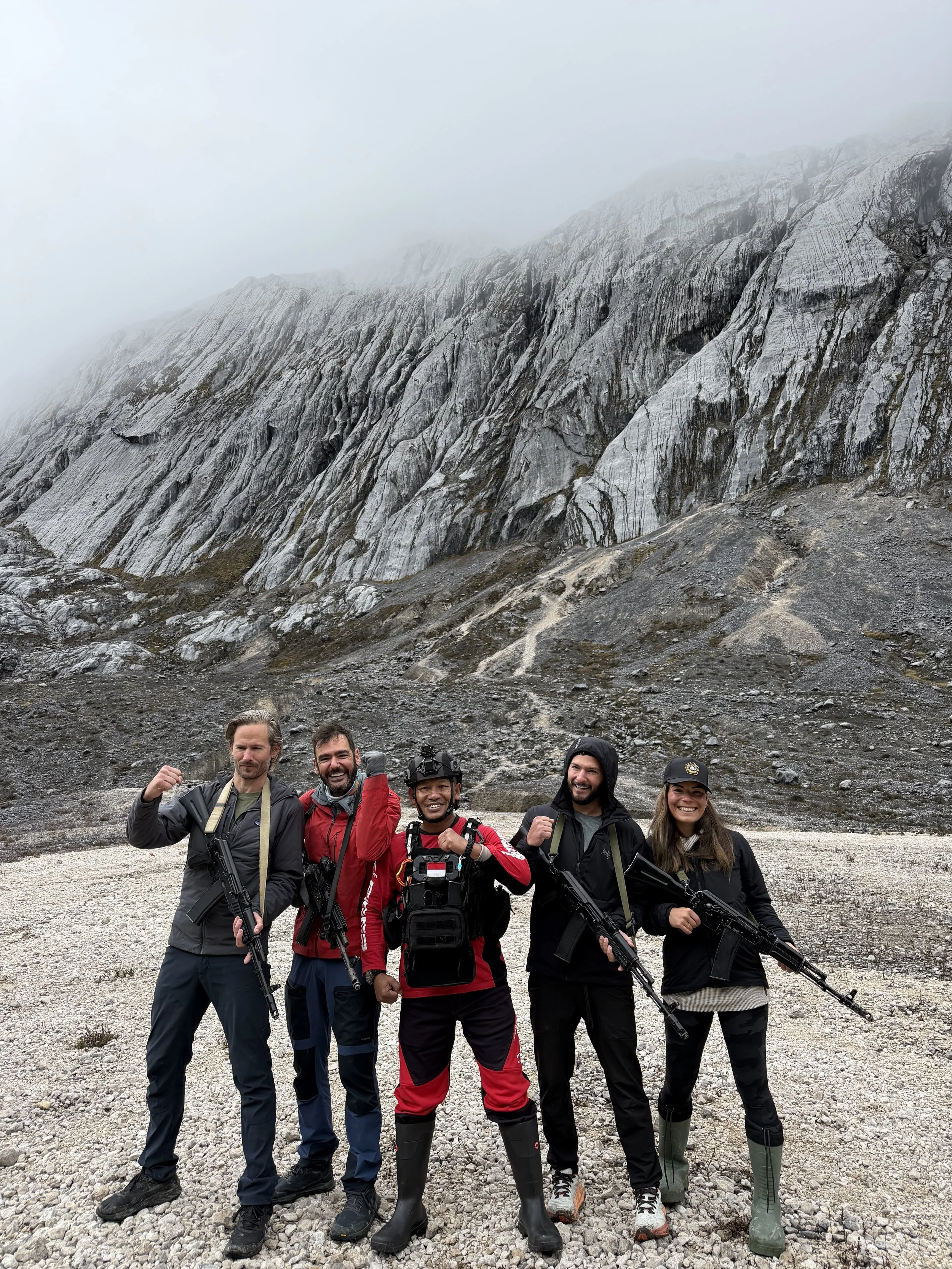 Group of five hikers with rifles posing in front of rocky mountain landscape with fog.