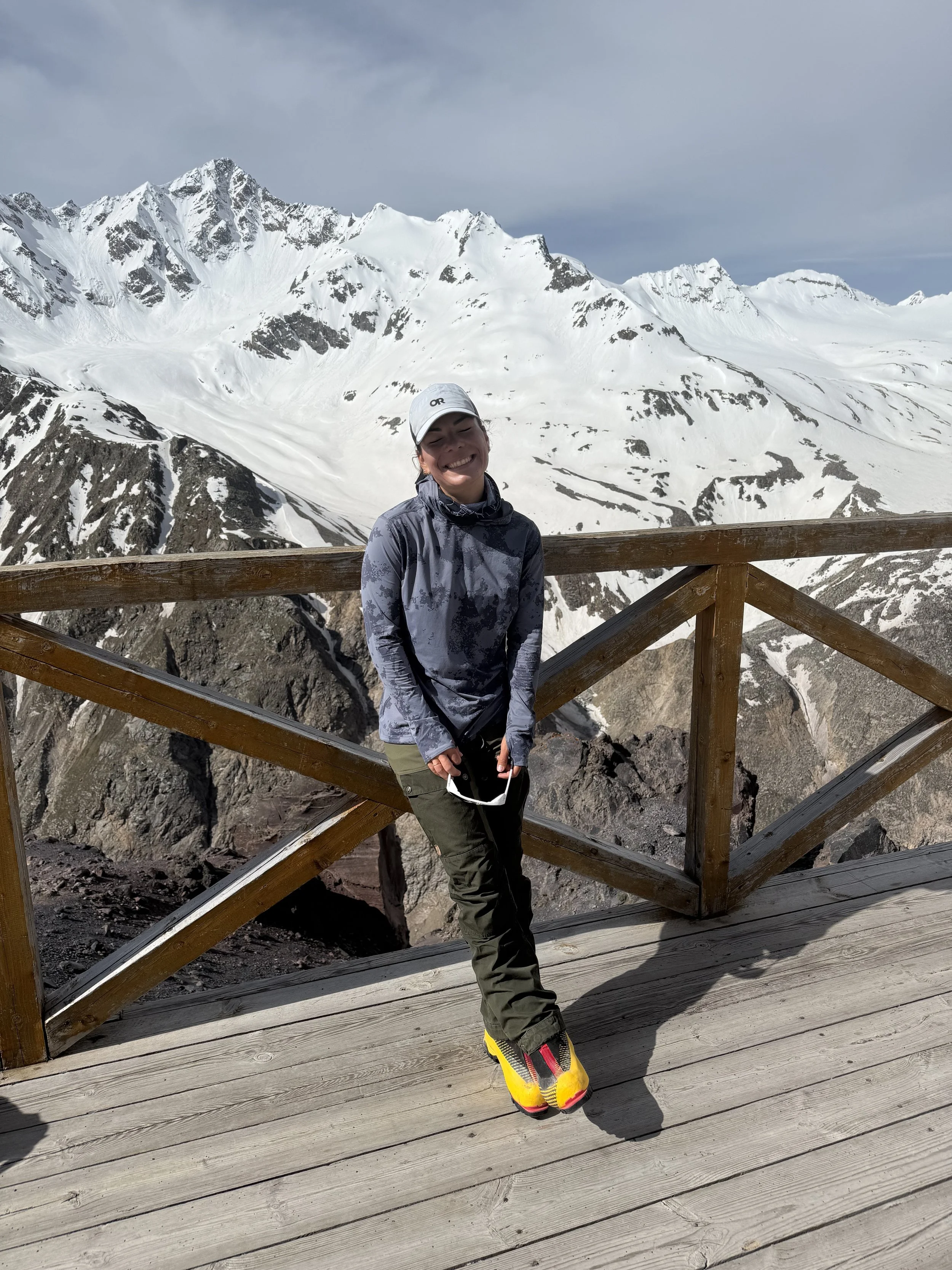 A woman standing on a wooden observation deck with snow-covered mountains in the background, wearing a cap, gray jacket, green pants, and yellow climbing shoes, smiling.