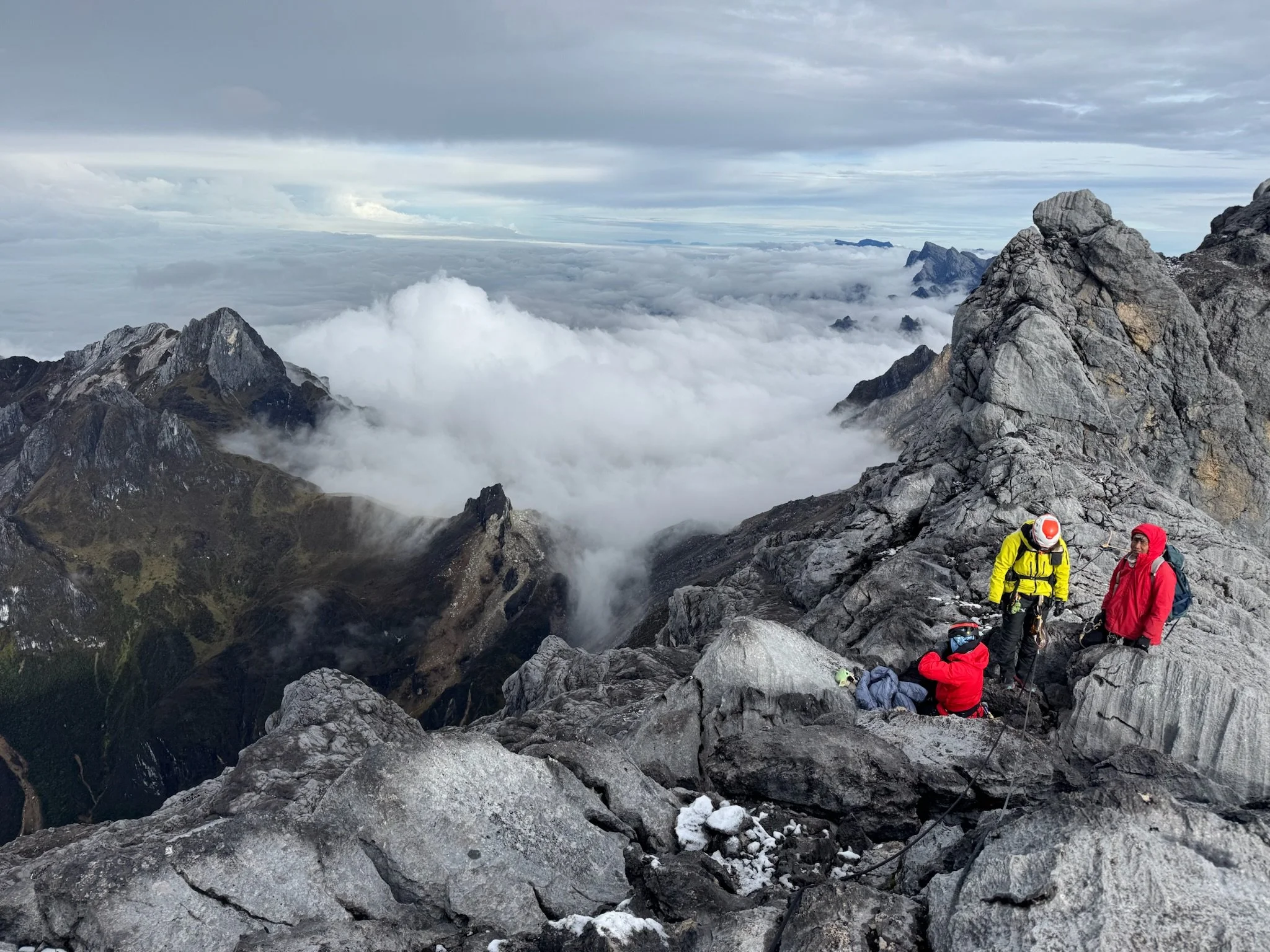 Three climbers in bright-colored jackets and helmets on a rocky mountain ridge, with clouds and distant mountain peaks in the background.