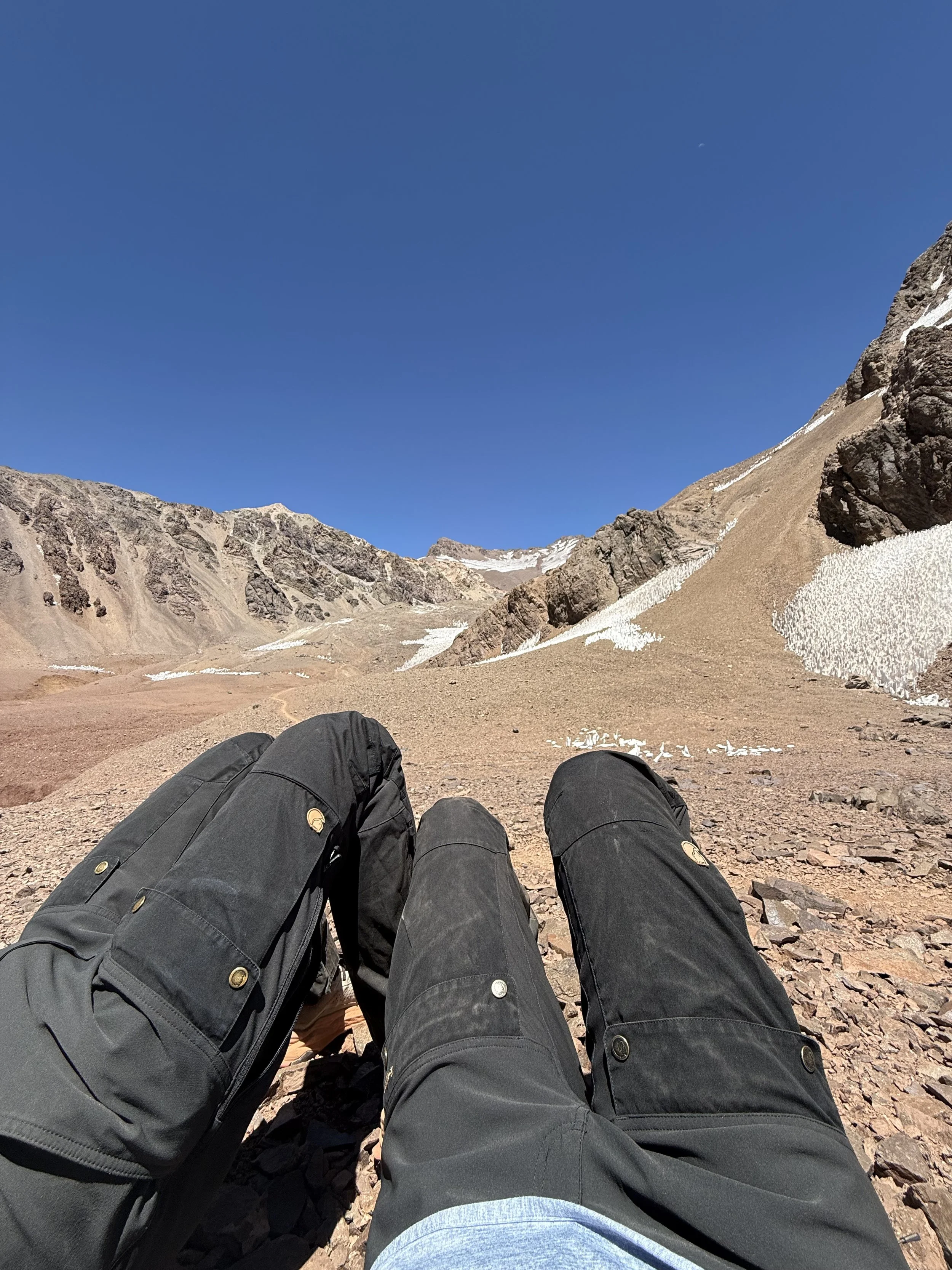 Two people with black backpacks and gray pants sitting on a rocky mountain terrain, looking at snow patches and rugged mountain peaks under a clear blue sky.